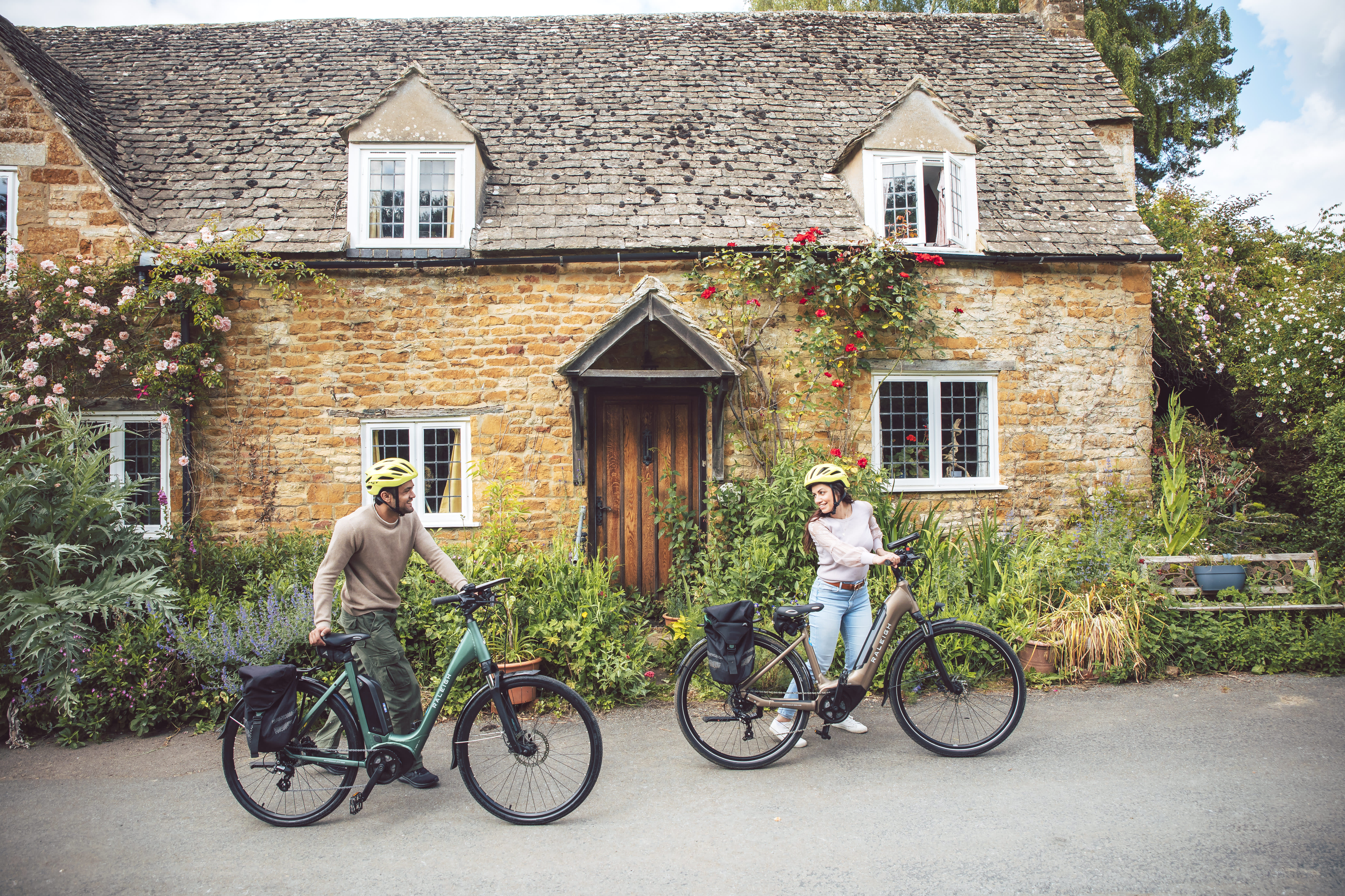 A couple push their bikes past a beautiful old cottage in a Cotswold village