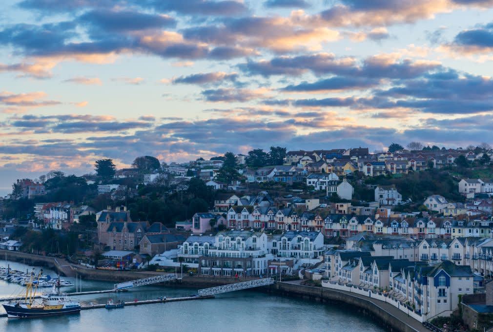 Coastal townscape at sunset, featuring colourful houses on a hillside, a calm marina with boats, and a cloud-streaked sky with warm hues. Tranquil ambience.