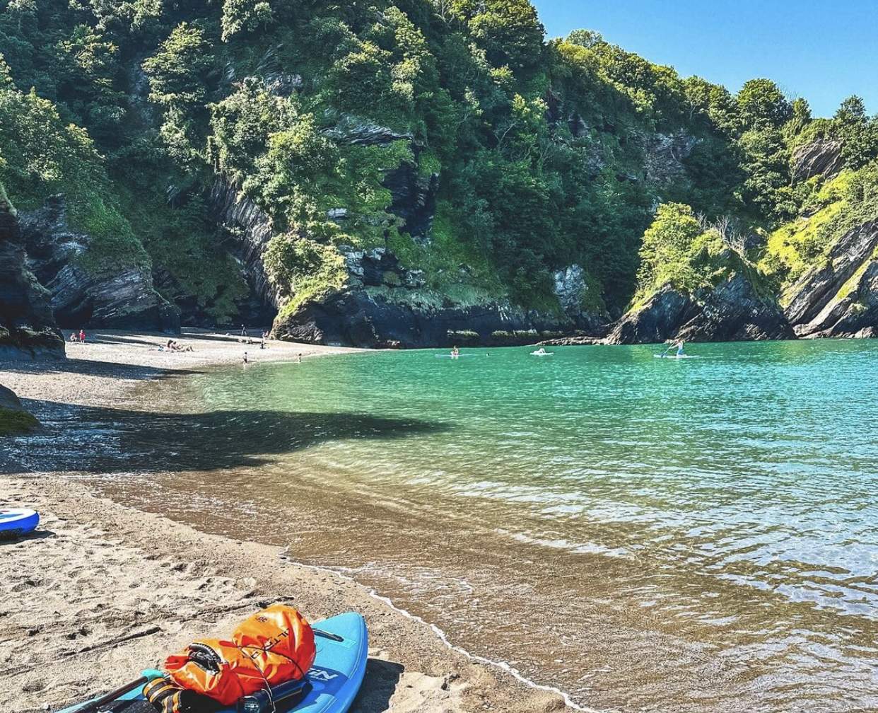 A serene cove with turquoise waters, surrounded by lush green cliffs. People are kayaking and relaxing on the sandy shore under a clear blue sky.