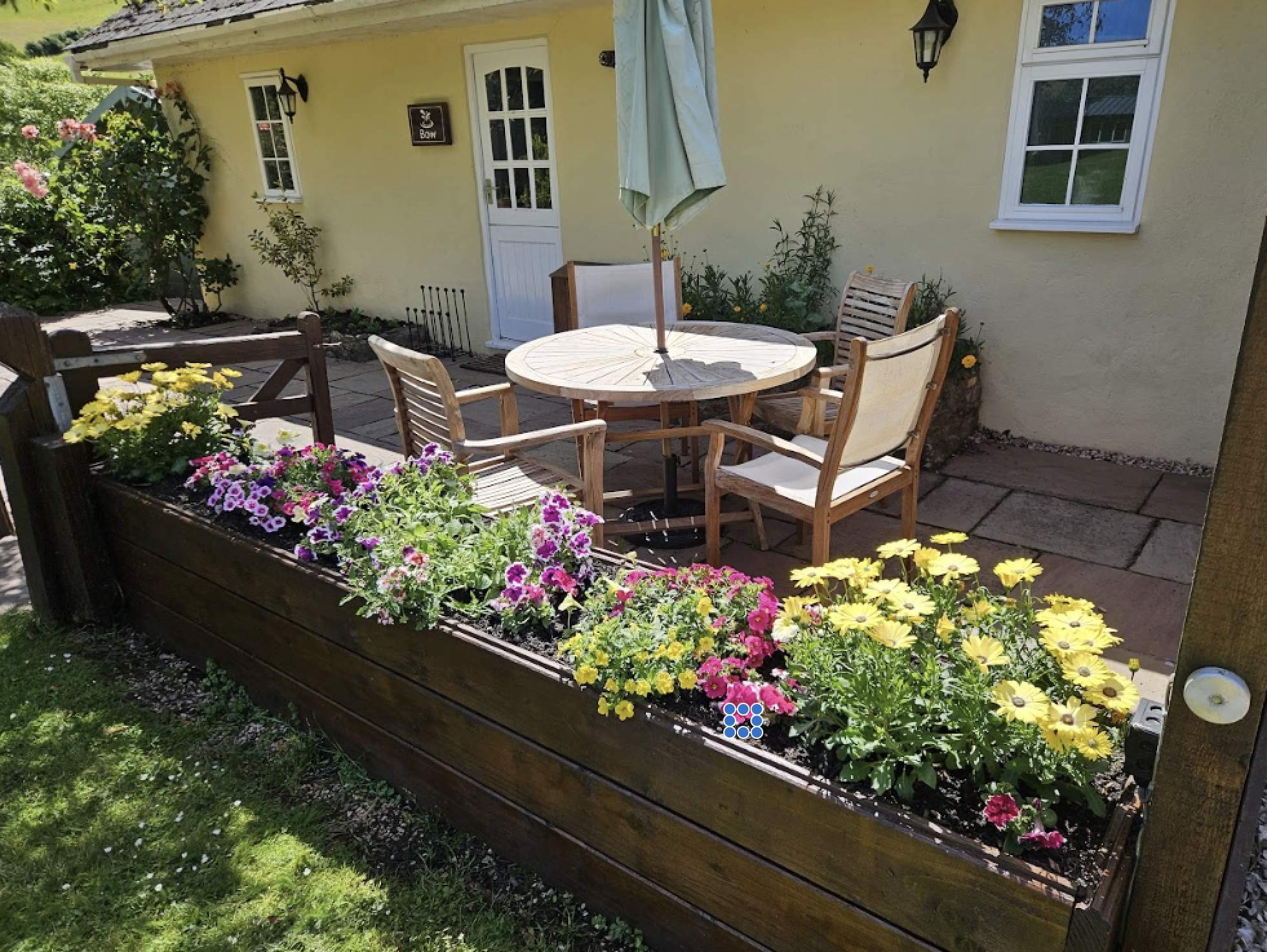 A cosy patio with wooden chairs and a round table shaded by a green umbrella. Vibrant yellow and pink flowers in a raised bed, backed by a yellow cottage.