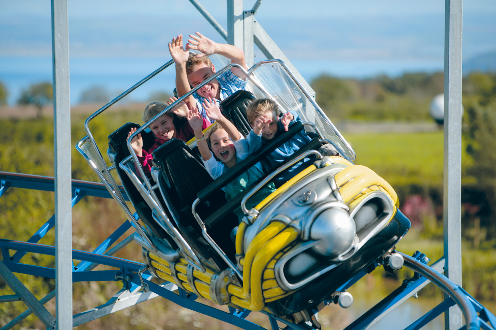 A group of excited people, including children, are enjoying a roller coaster ride. They're smiling, hands raised, surrounded by a sunny, green landscape.