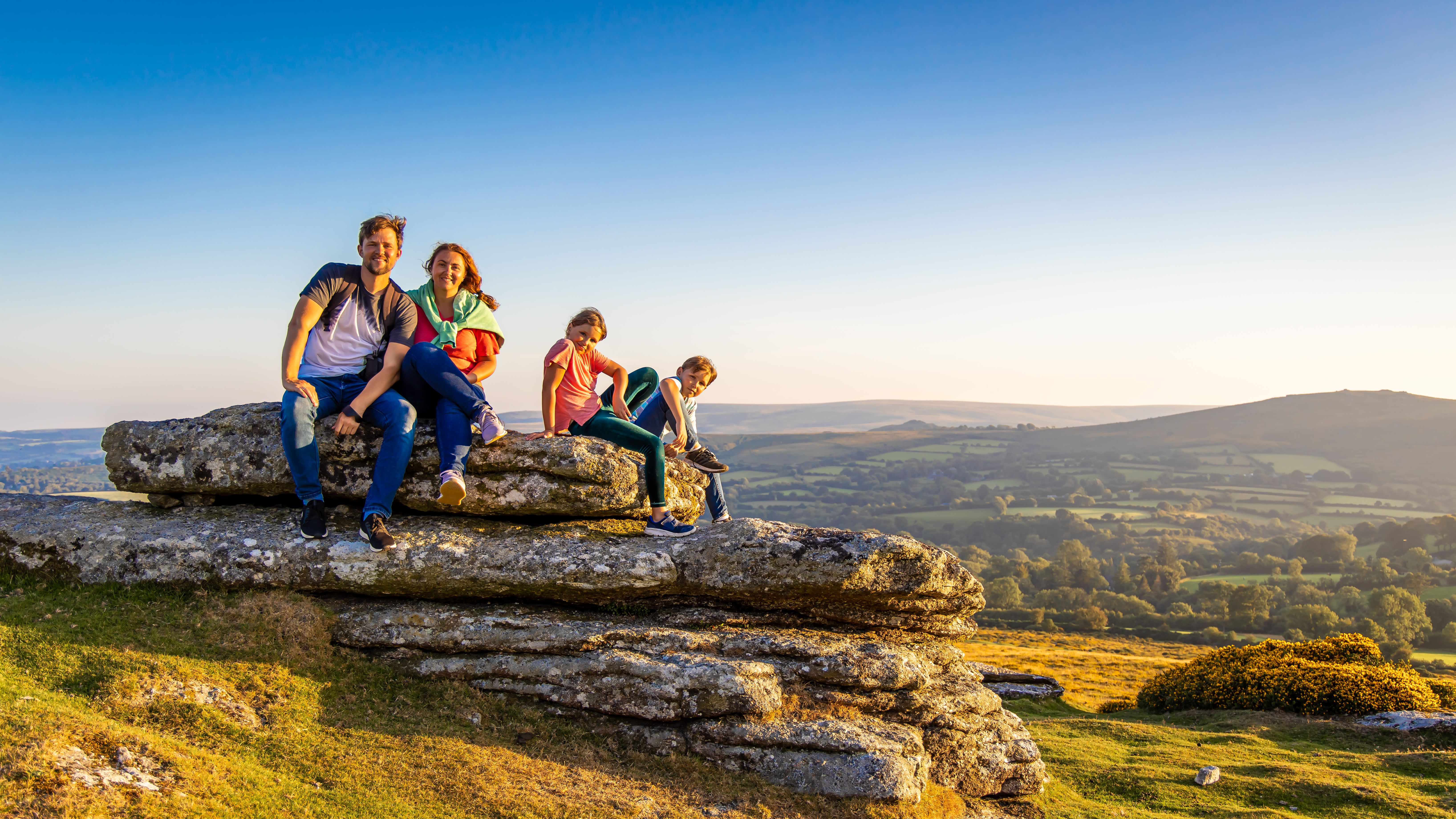 a family of two adults and two children sitting on a flat rock in Dartmoor with spectacular view behind them