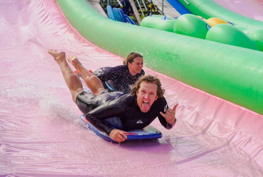 Two people in wetsuits joyfully slide down a vibrant pink water slide on bodyboards. The front person makes a playful face, conveying excitemet.