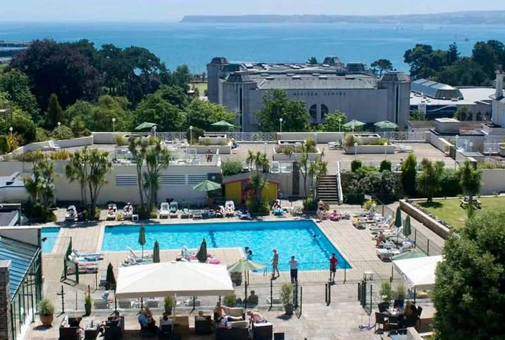 Aerial view of a vibrant hotel pool area with people lounging under umbrellas and swimming, surrounded by lush greenery and overlooking a serene ocean.