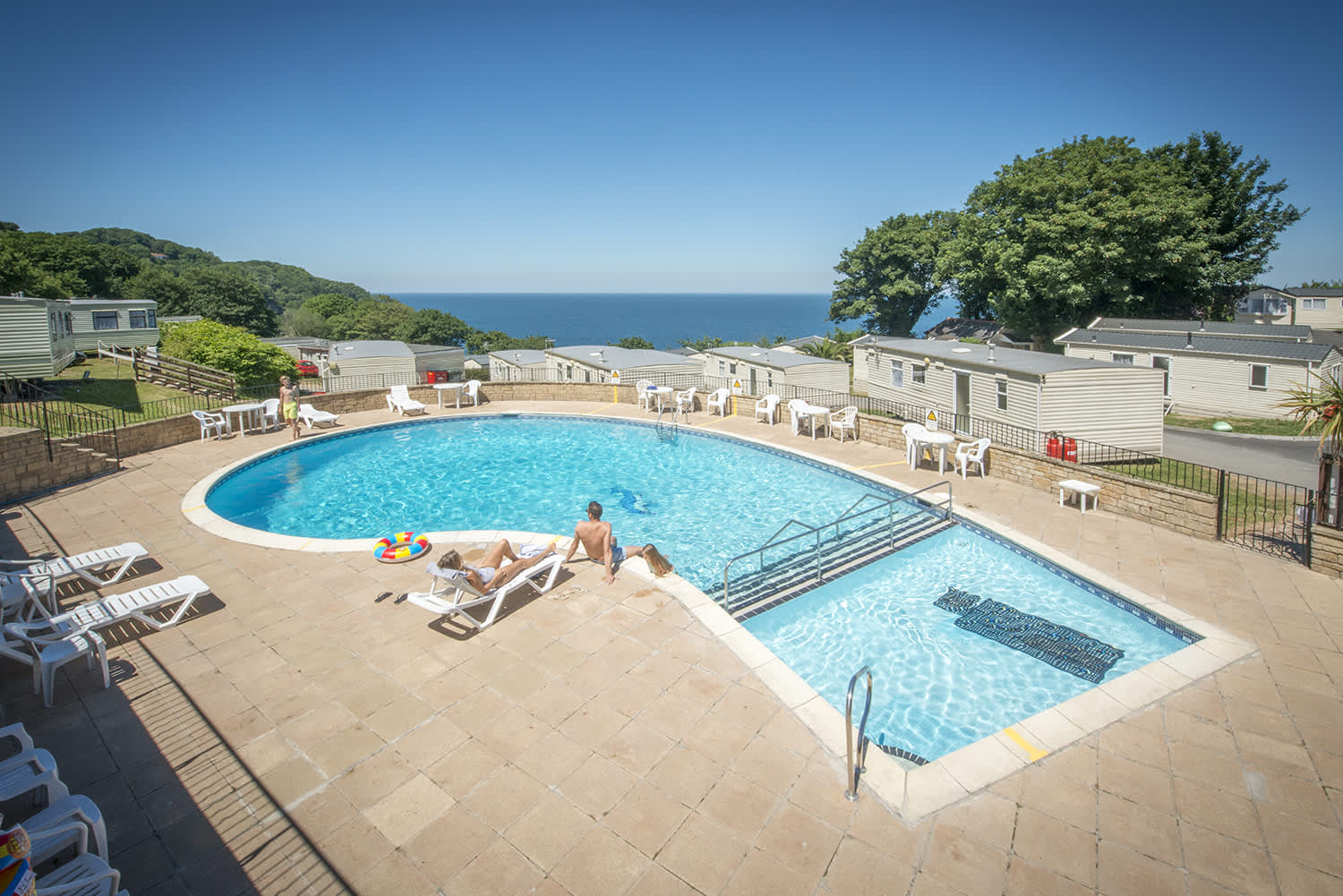 Outdoor pool with loungers, people sunbathing, and a colourful inflatable under a clear blue sky. Mobile homes and sea view in the background.