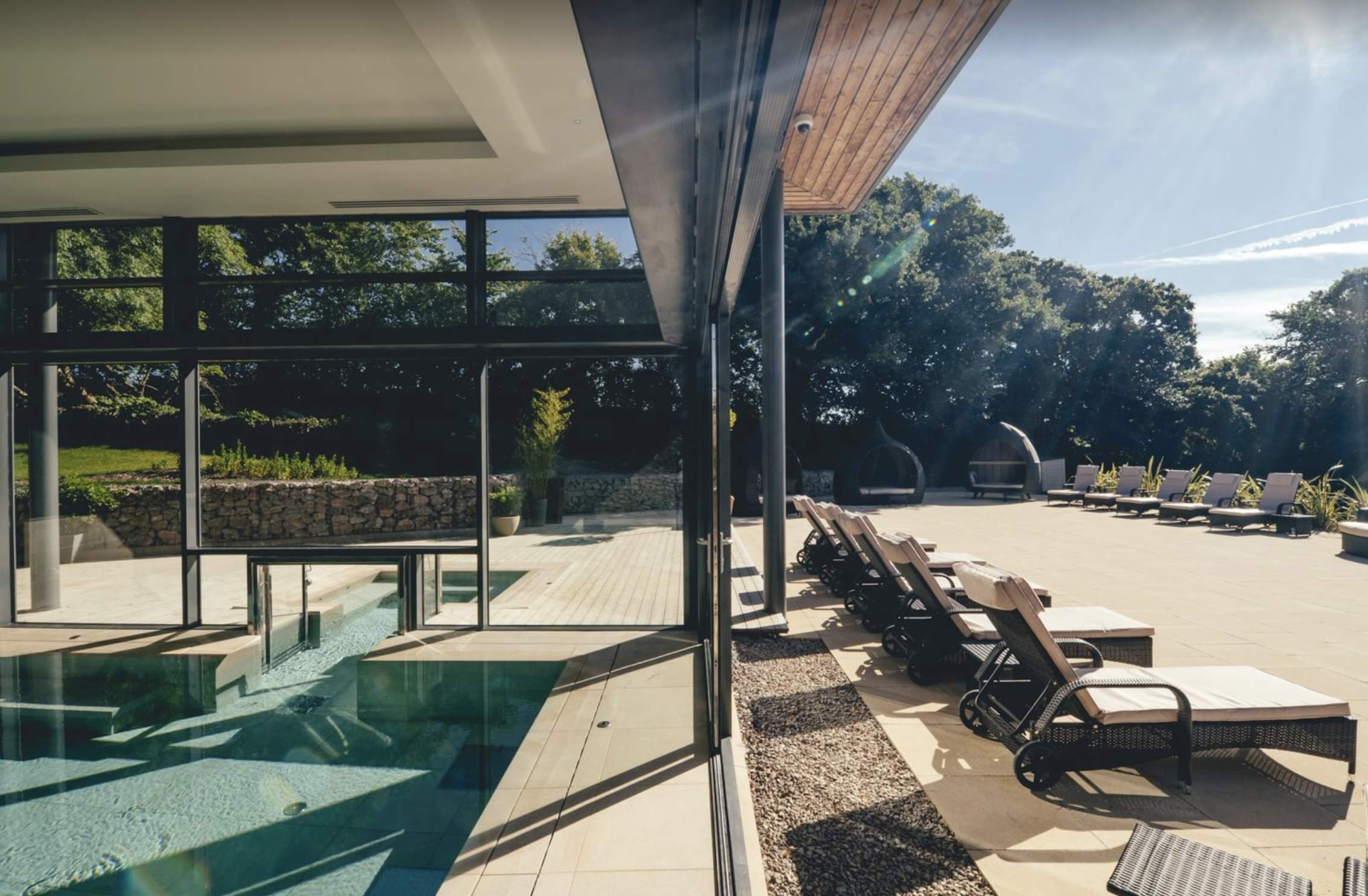 Luxurious pool area with elegant lounge chairs on a sunlit deck. Glass walls separate indoor pool from outdoor space, surrounded by lush greenery.