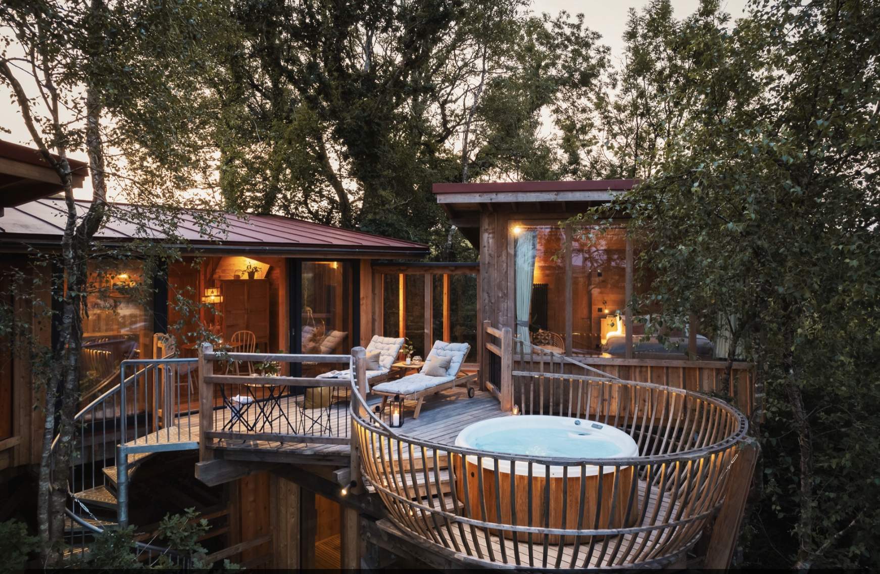 Treehouse retreat at sunset with large windows, cosy interior lighting, chaise lounges, and a hot tub on a wooden deck surrounded by lush trees.