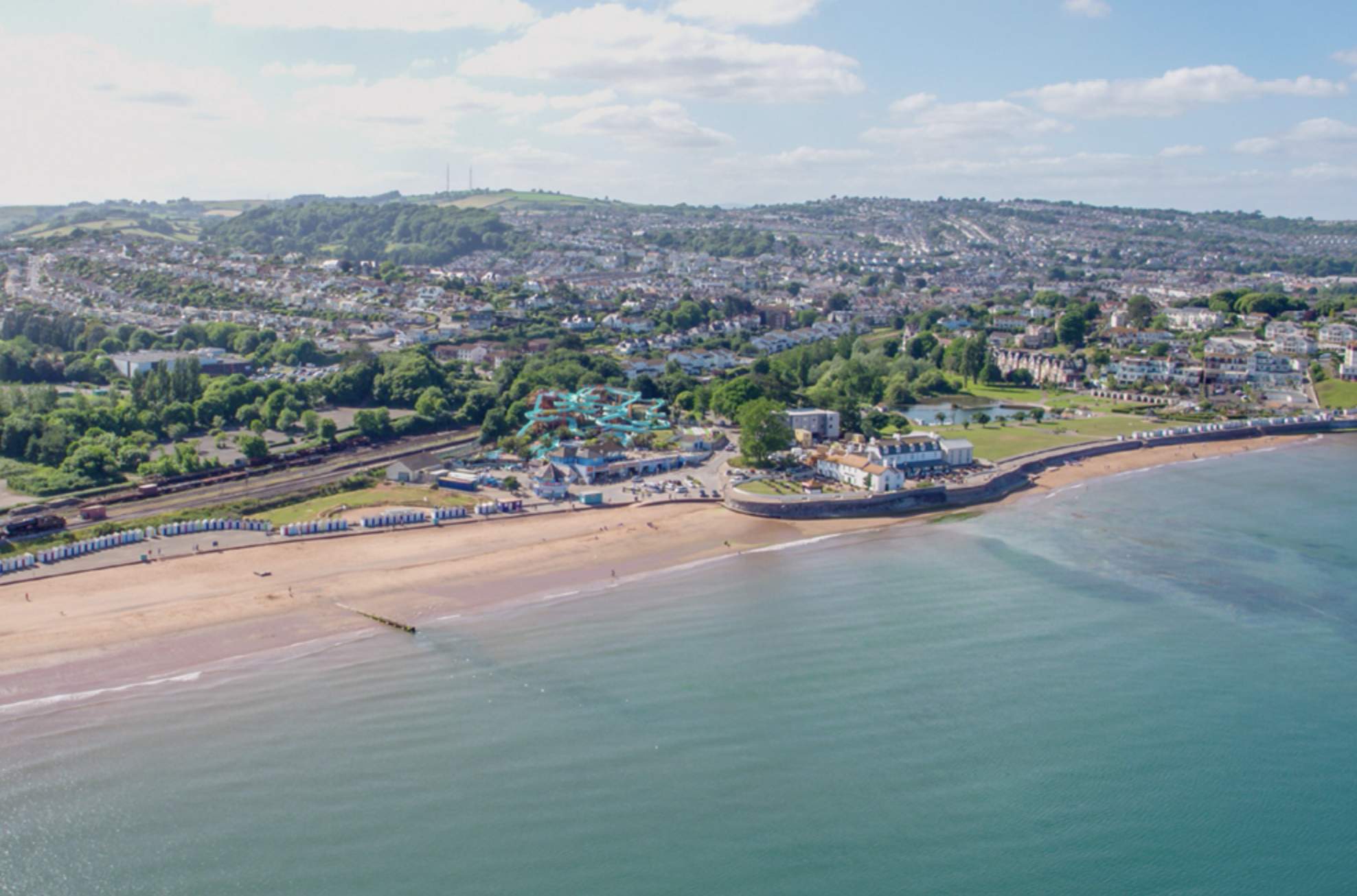 Aerial view of a coastal town with a sandy beach lined with blue beach huts. The sea is calm, and a scenic hillside town is in the background. The tone is peaceful.