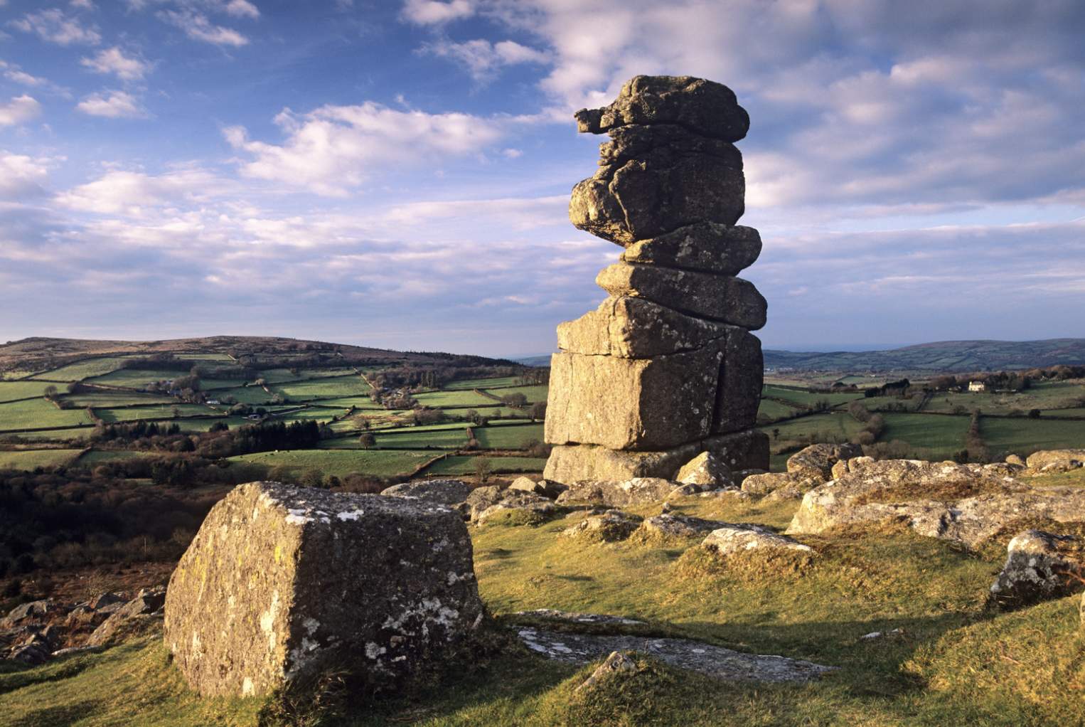 Towering rock formation on Dartmoor overlooks rolling green fields under a blue sky. Sunlit stones create a serene, ancient atmosphere.