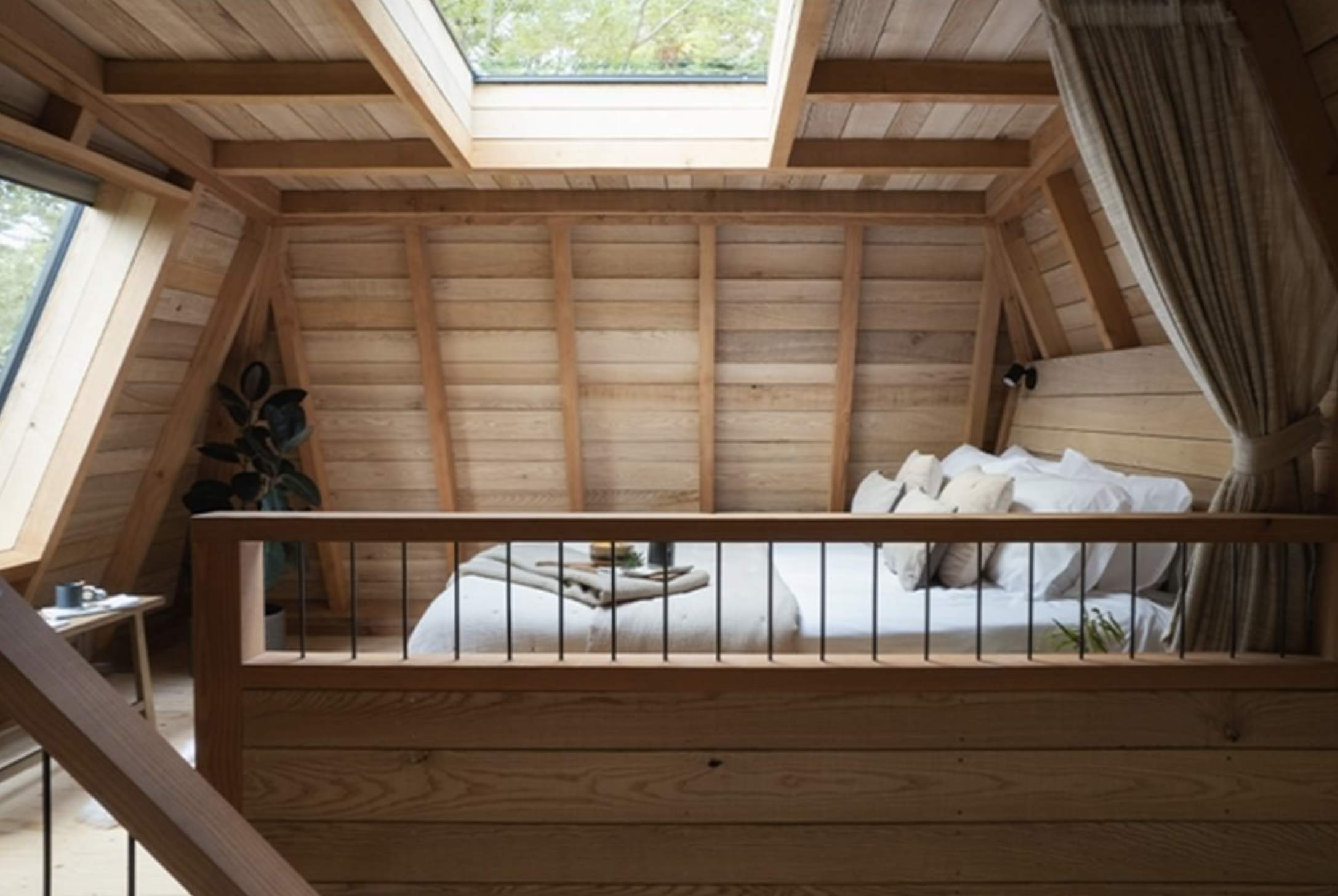 A cosy attic bedroom with wooden walls and ceiling, featuring a skylight above a neatly made bed with cushions. Soft natural light creates a warm ambiance.