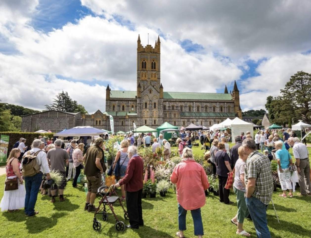 Crowded outdoor garden festival in front of a large, historic cathedral. People browse plant stalls under partly cloudy skies, conveying a lively atmosphere.