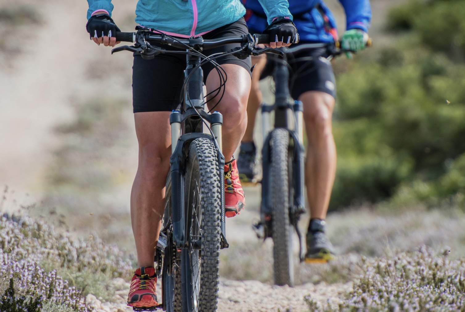 Two people riding mountain bikes on a rocky trail. They wear colourful jackets, shorts, and gloves. The background is blurred with greenery, conveying motion and adventure.