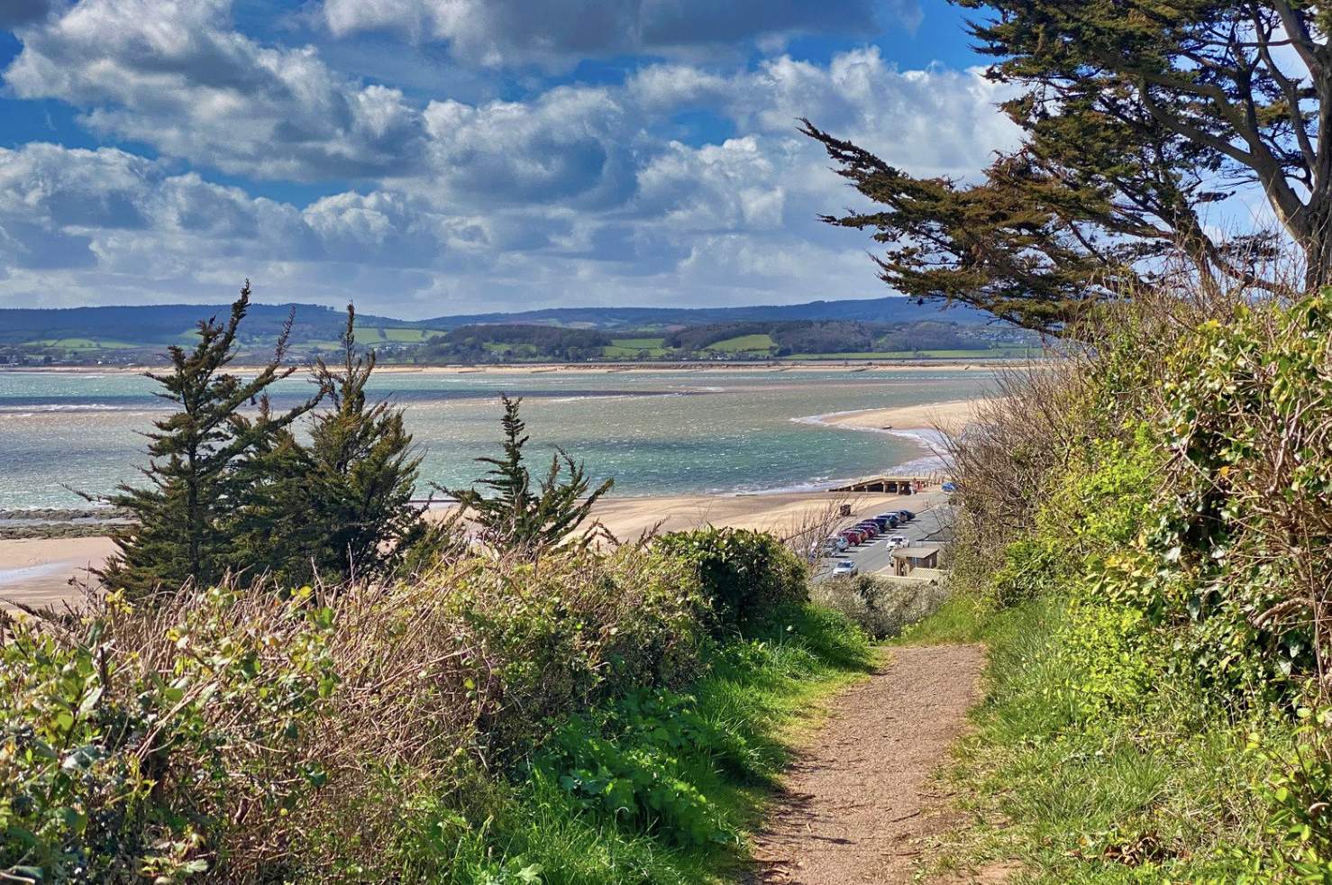 A scenic coastal path leads to a sandy beach, bordered by lush greenery and trees. The sea sparkles under a partly cloudy sky, conveying a serene atmosphere.