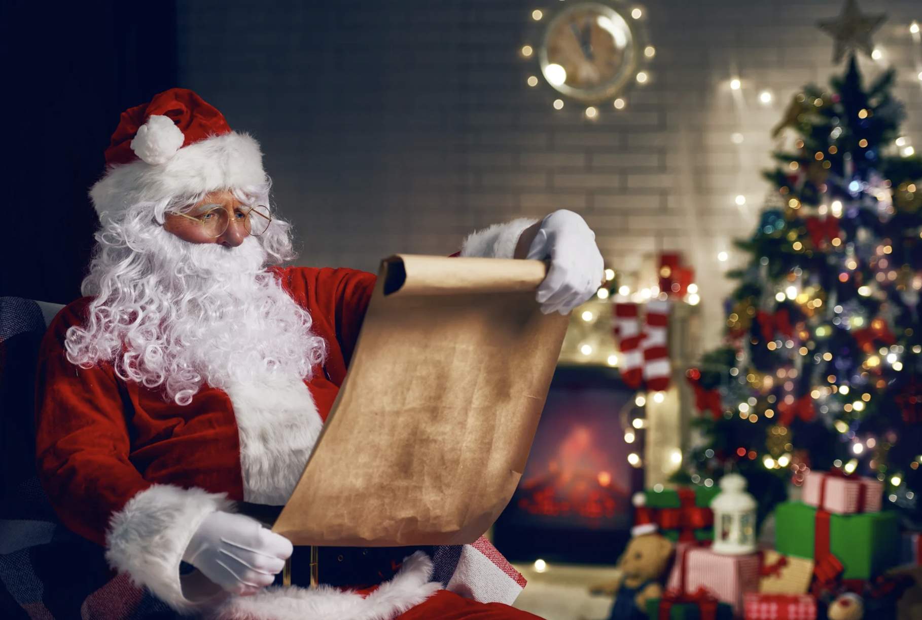 Santa Claus in a red suit reads a scroll by a cozy fireplace. A decorated Christmas tree and wrapped gifts are in the background, creating a festive atmosphere.