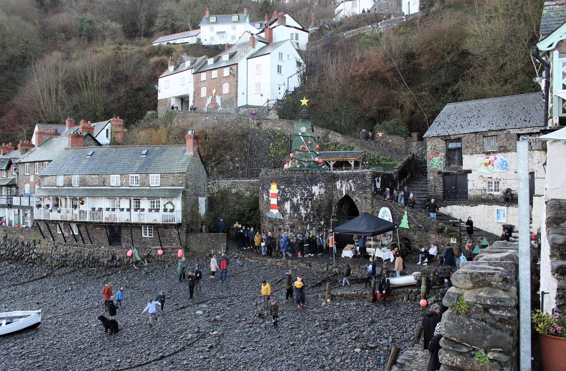 Coastal village scene with festive decorations. People gather near stone buildings and a Christmas tree. Pebbled shore in foreground and hillside houses behind.
