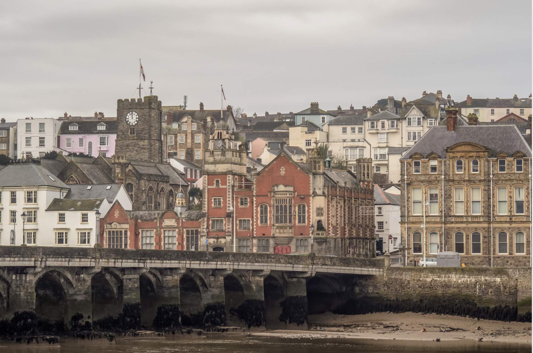 Historic townscape featuring a stone bridge, Victorian buildings, and a clock tower. The overcast sky and muted colors create a nostalgic atmosphere.