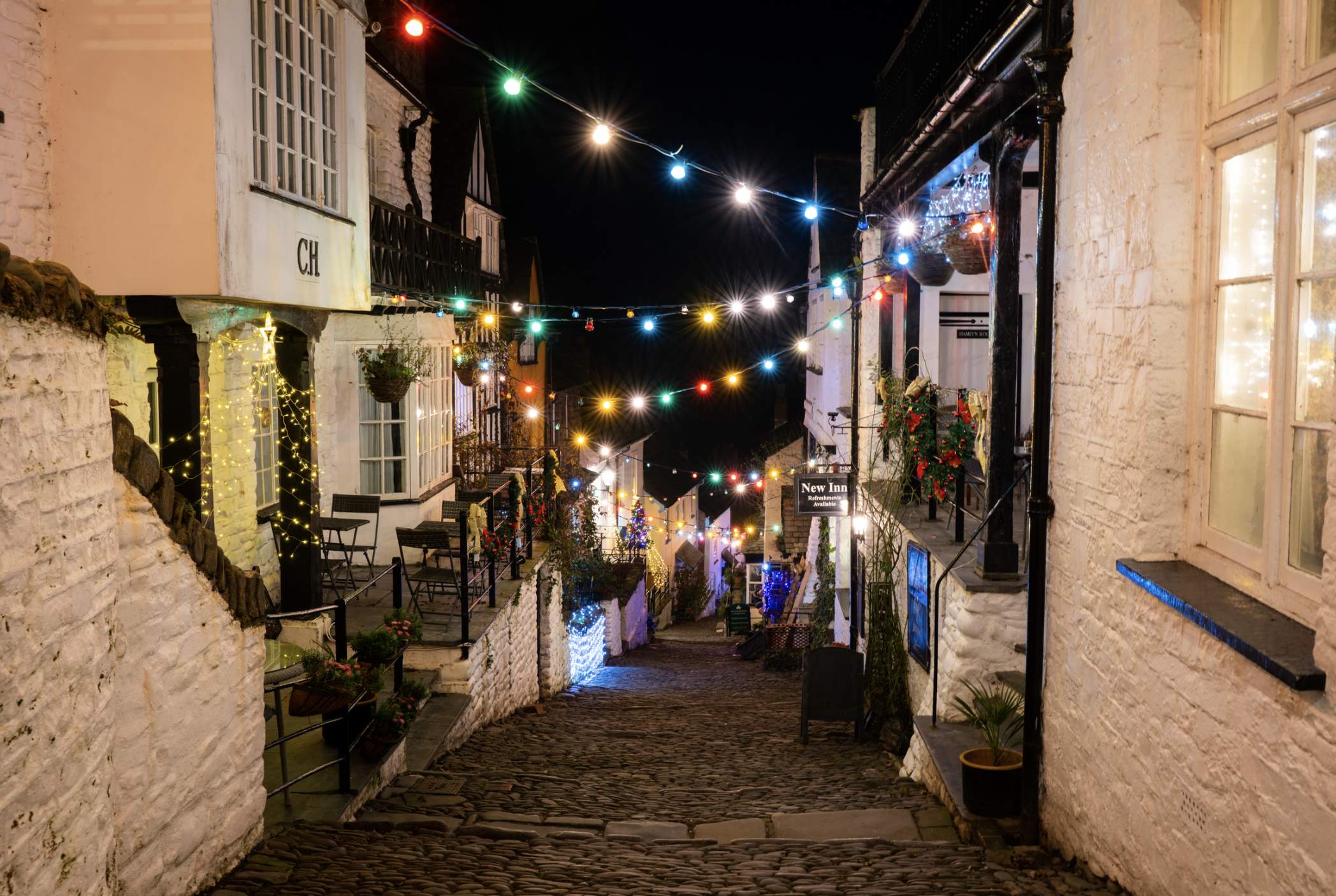 A charming cobblestone street at night, lined with cosy houses adorned with glowing multicoloured string lights, creating a festive, warm atmosphere.
