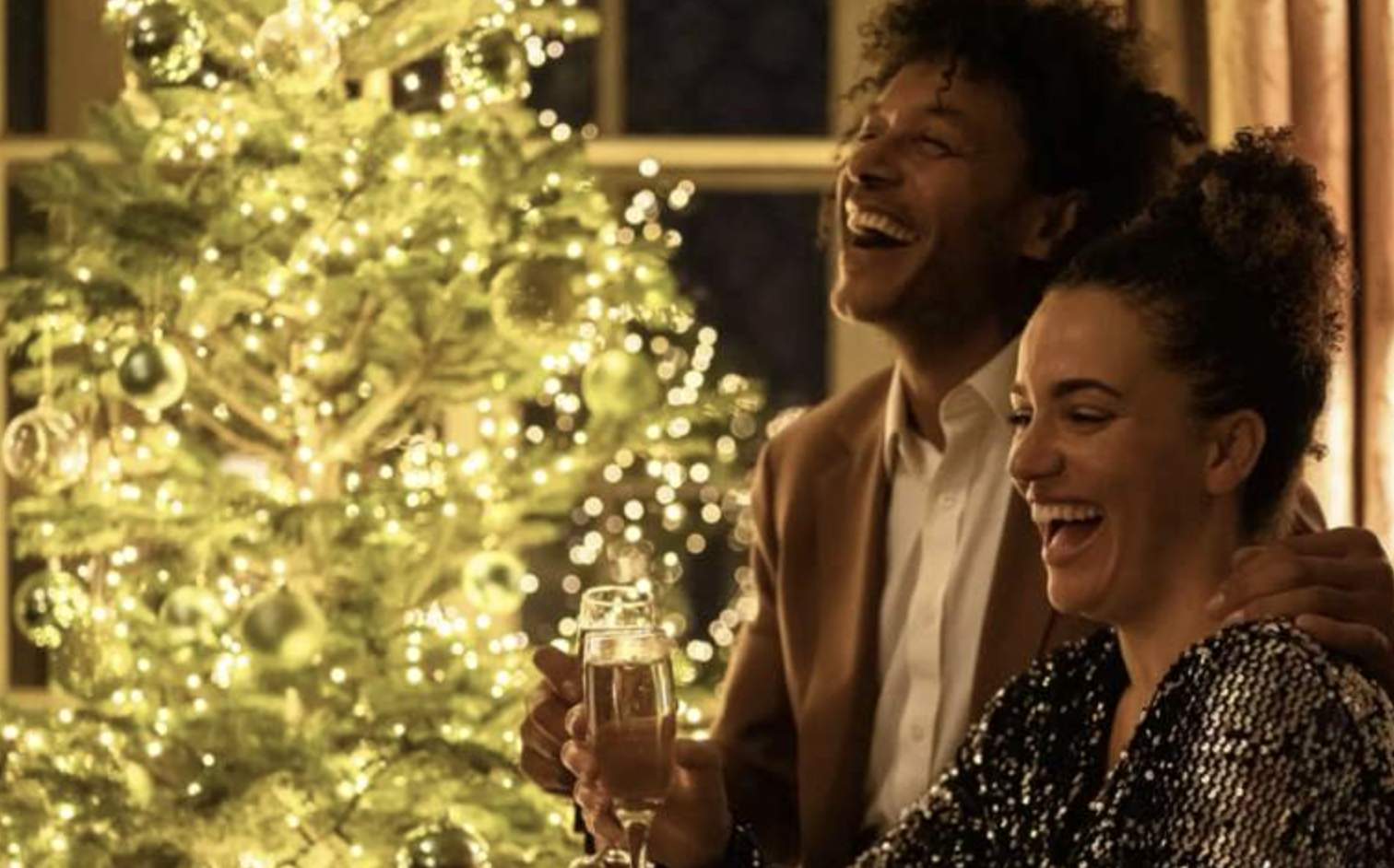 A joyful couple celebrates near a sparkling Christmas tree at Mole Resort holding champagne glasses and laughing warmly. The room glows with a festive and cosy ambience.