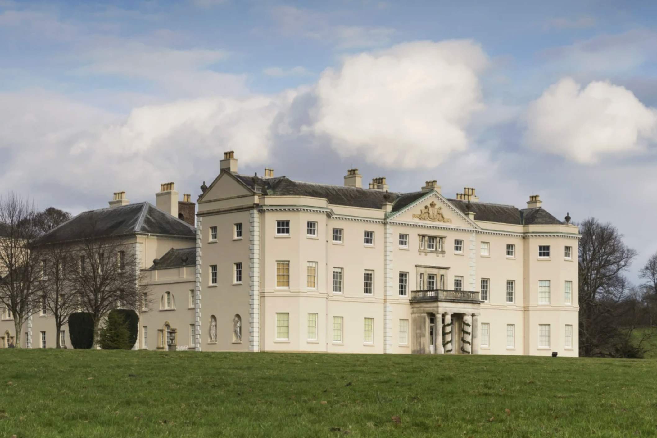 A grand historic Saltram House, cream-colored walls and tall windows set against a blue sky with fluffy clouds, surrounded by green lawns and leafless trees.