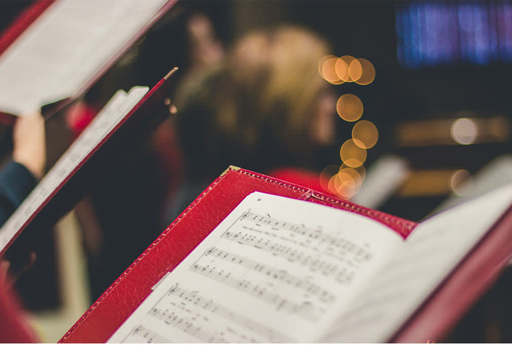 Close-up of sheet music in red folders held by choir singers, blurred background with warm bokeh lights, conveying a festive and serene atmosphere.