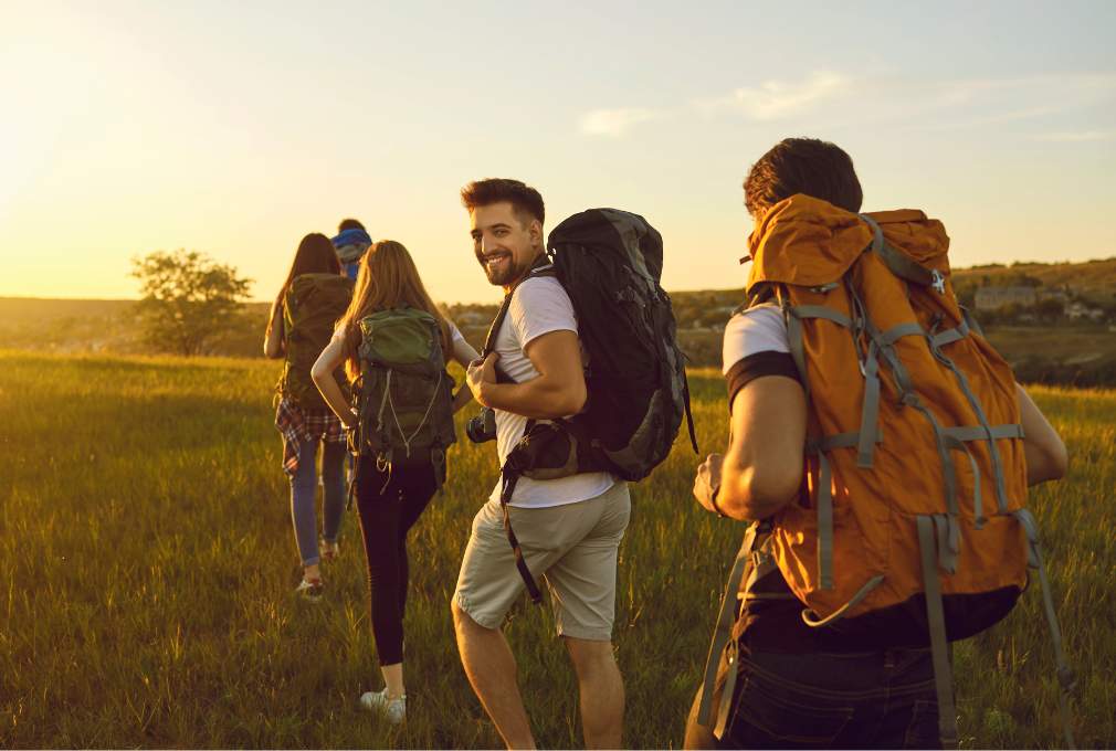 Four hikers with backpacks walk through a field at sunset. One glances back smiling, conveying a sense of adventure.
