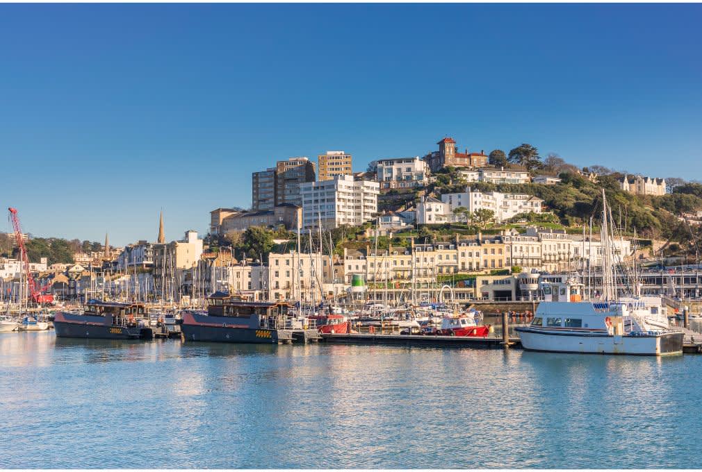 Coastal townscape with boats docked in a marina, set against a hillside of buildings under a clear blue sky, conveying a serene, leisurely vibe.