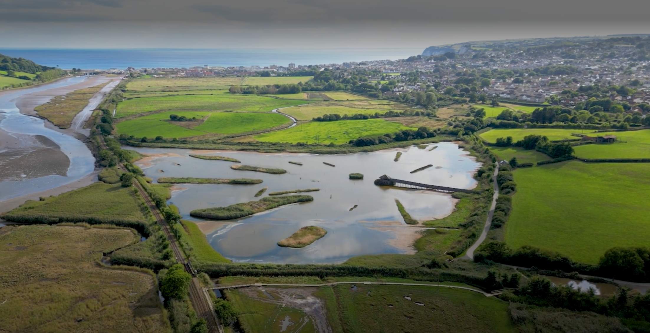 Aerial view of a scenic wetland area with ponds, surrounded by vibrant green fields and distant hills. A tranquil landscape under a clear sky.