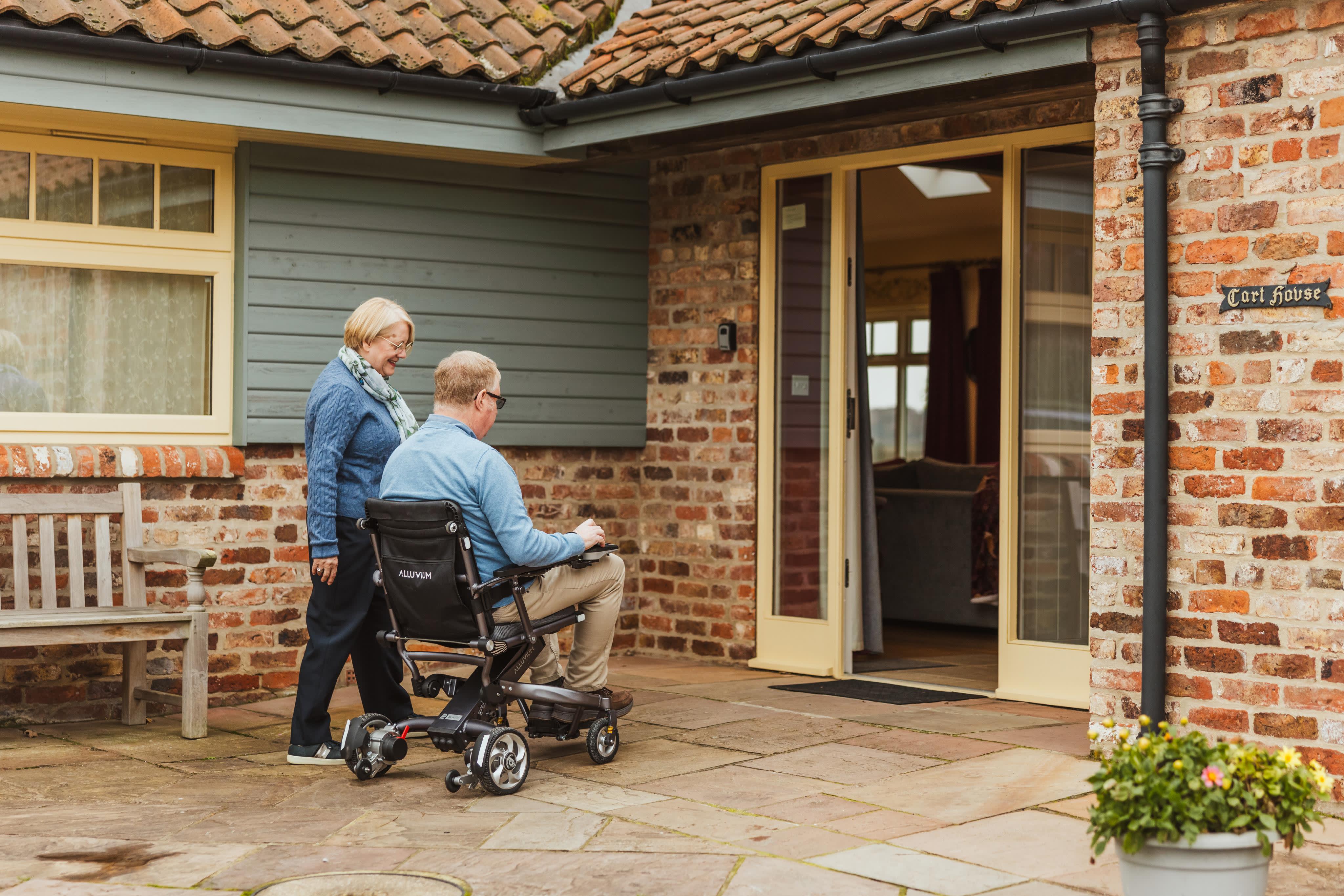 2 people 1 in motorized wheelchair approaching a cottage door
