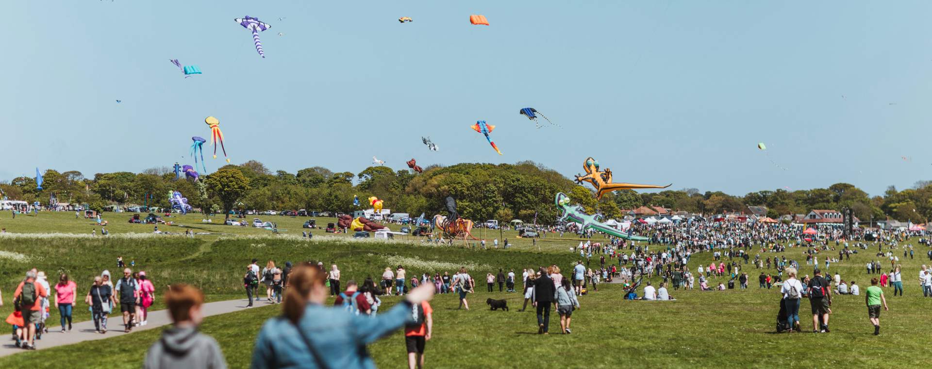 A large crowd walking towards the Bridlington Kite Festival, with kites floating in the air in the distance