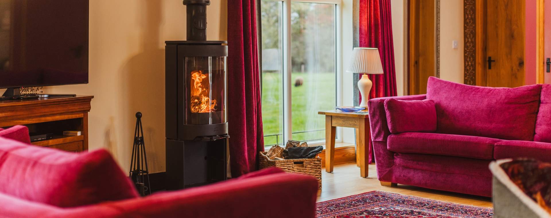The inside of a cosy cottage, with a log burning stove and two magenta sofas.