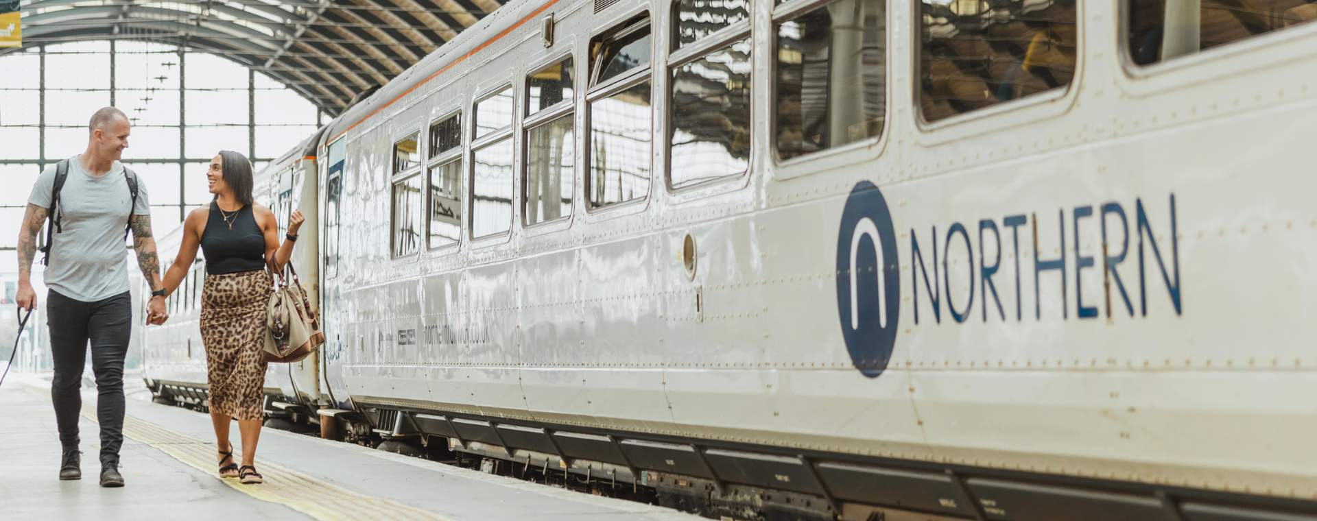 A couple walking hand-in-hand along a train station platform. A Northern train is stationary on the tracks beside them.