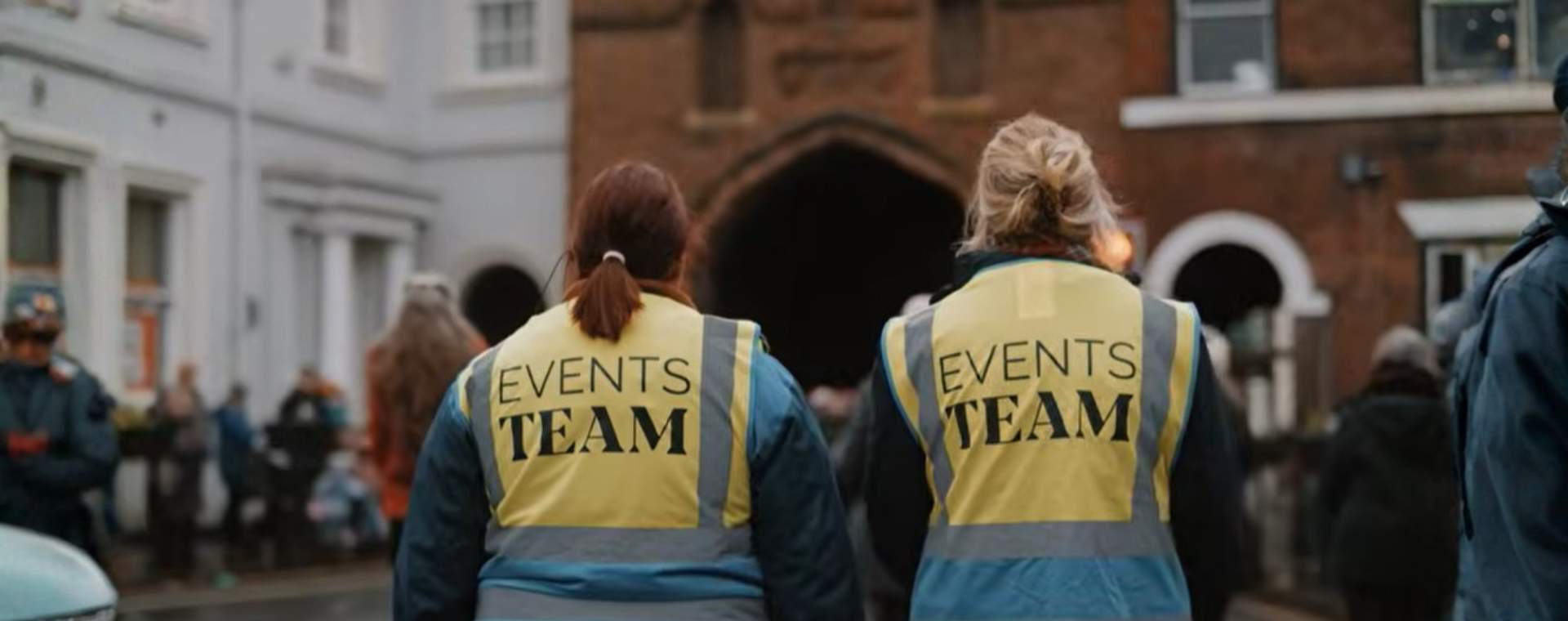 The back of two people walking towards North Bar, wearing hi vis jackets reading 'Events Team' on the back.