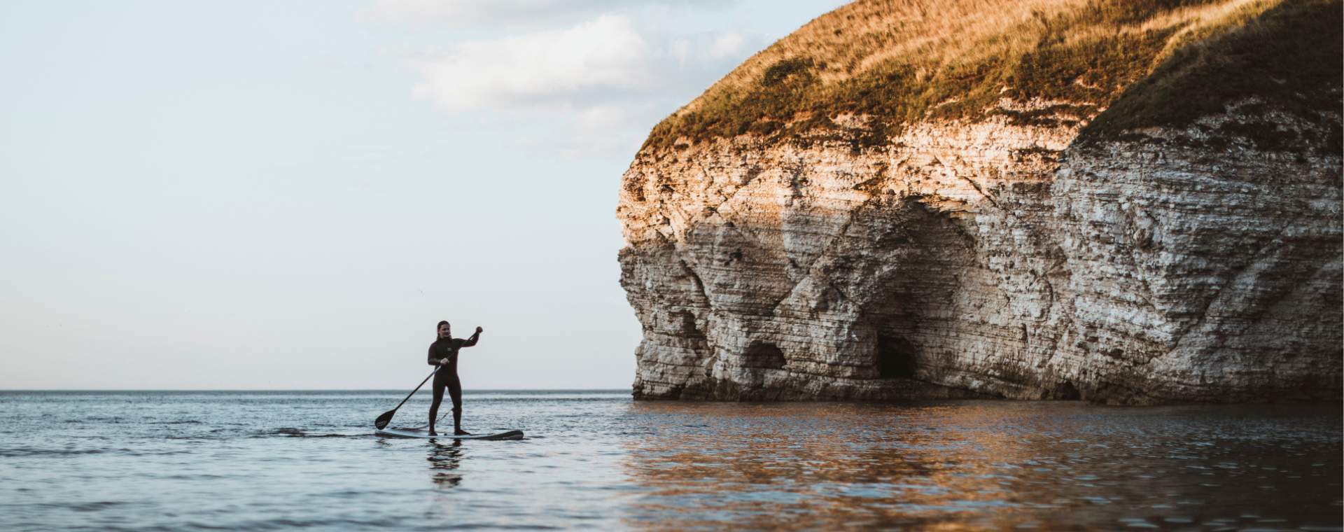 A man paddle boarding in the sea, with the sun appearing on the cliff edge at North Landing, Flamborough