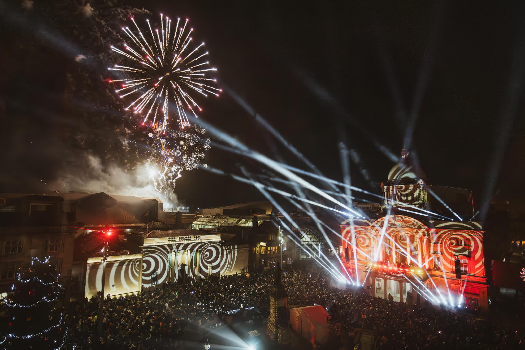 An overview of Hull City Hall and the Maritime Museum, lit up at night with spirals and flashlights, whilst fireworks explode overhead. A large crowd is gathered to watch the display.