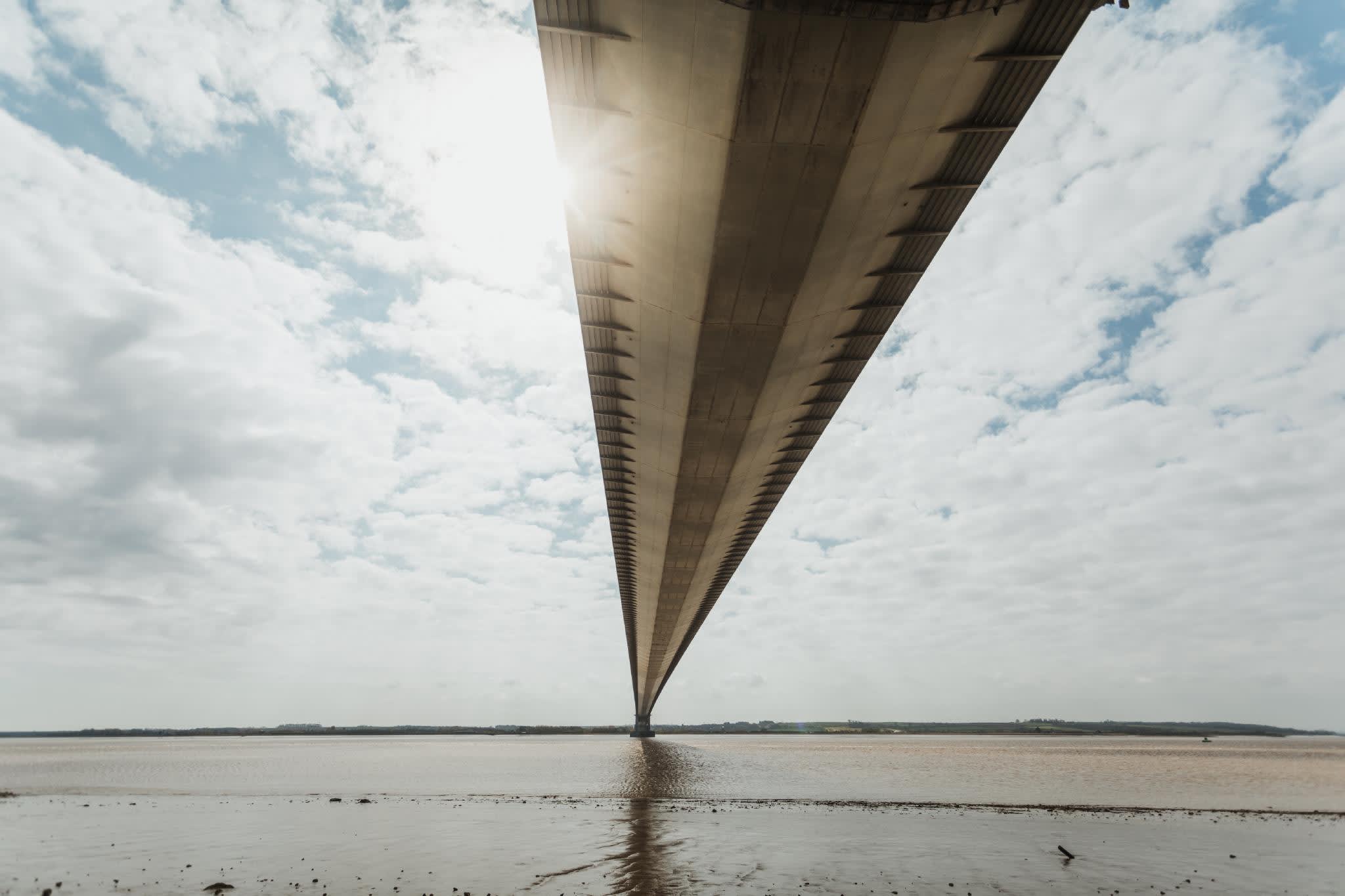 A view beneath the Humber bridge, with clouds either side and the foreshore just in front of the viewer.