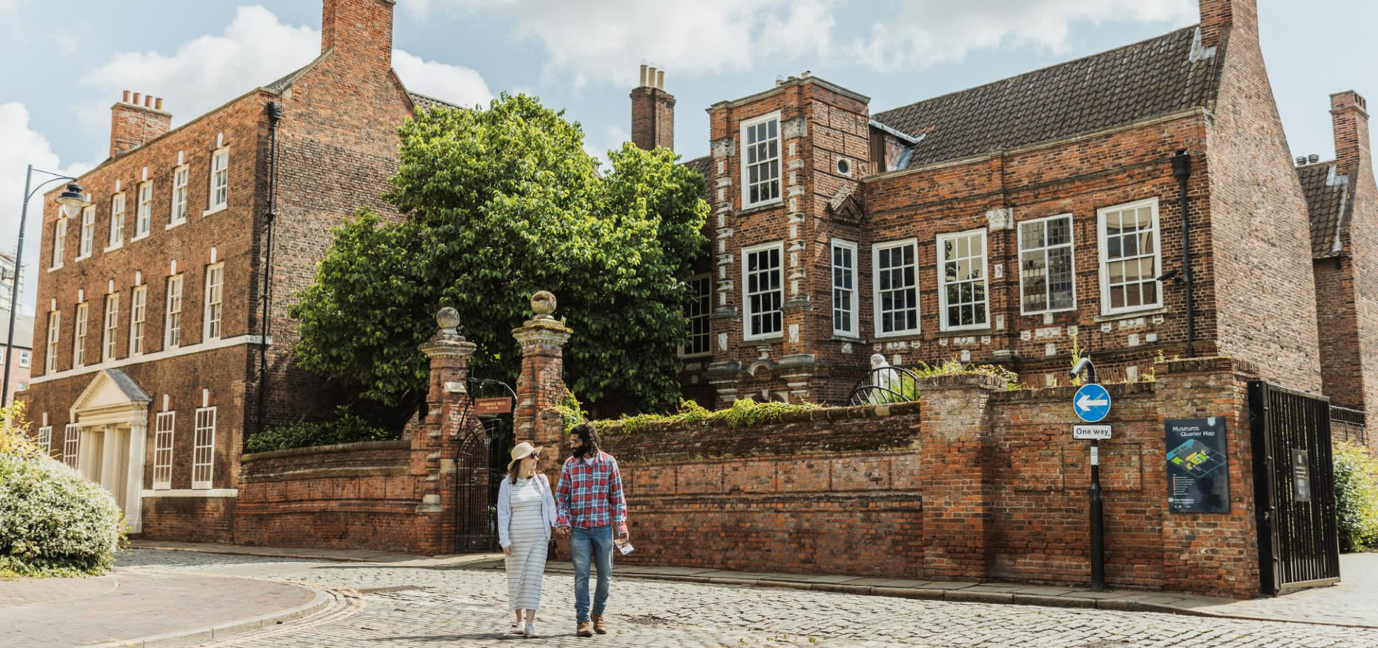 A cobbled street scene outside a historic red‑brick building in Hull’s Museums Quarter, with two visitors walking past the Wilberfoce House under a bright, clear sky.