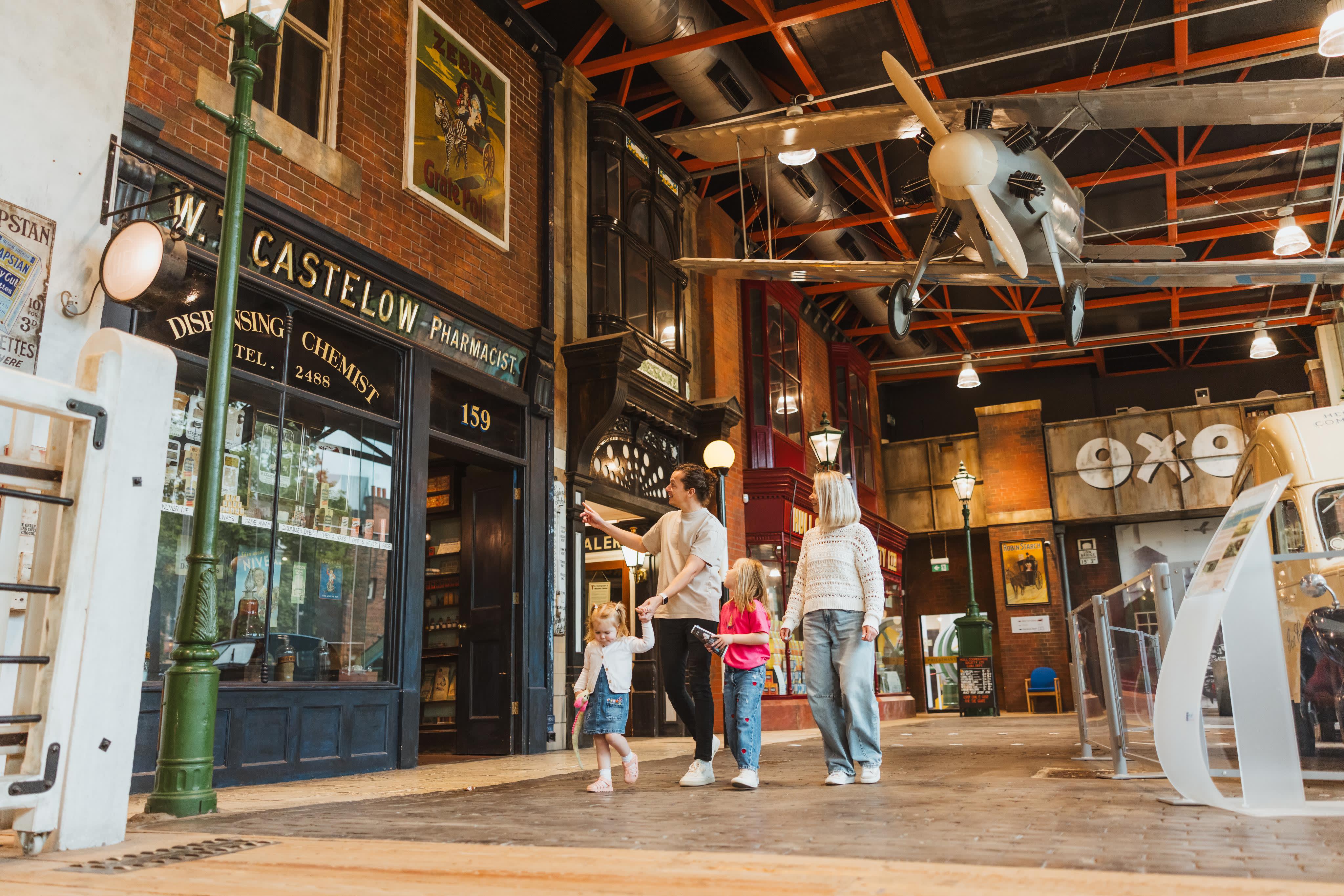 A family of 2 adults and 2 children looking in a museum. A vintage aeroplane is above their heads.