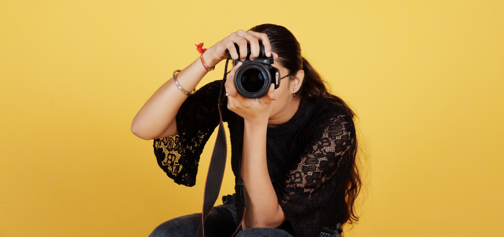 A woman holds a camera up covering her face in front of a yellow background