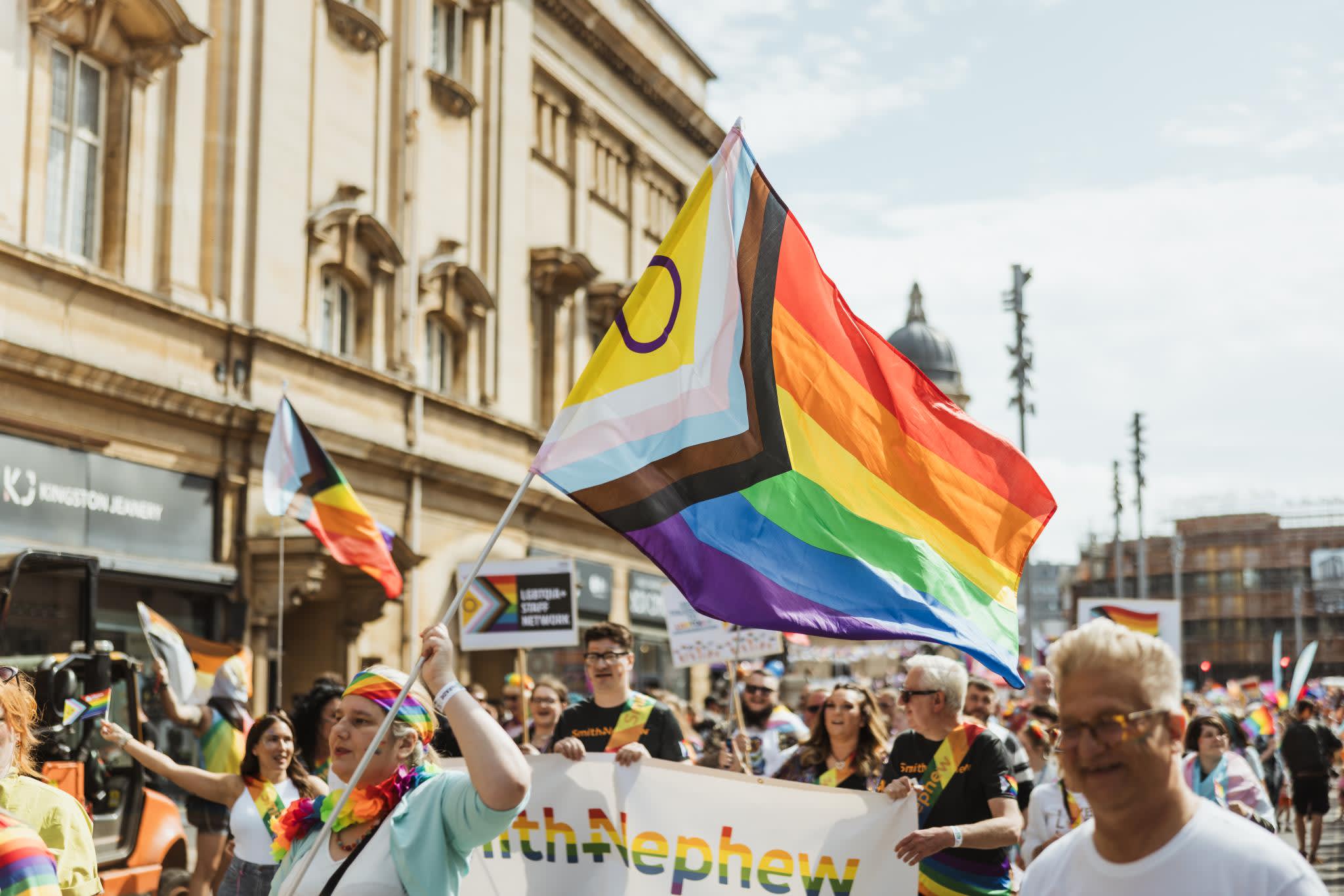Pride In Hull's parade walking down Carr Lane. The sun is shining, the crowd is smiling and dressed in shorts and t-shirts. A group of people holding a rainbow Smith+Nephew banner are in the foreground with a brightly dressed woman waving an Intersex Inclusive pride flag. Many other pride flags can be seen in the background.