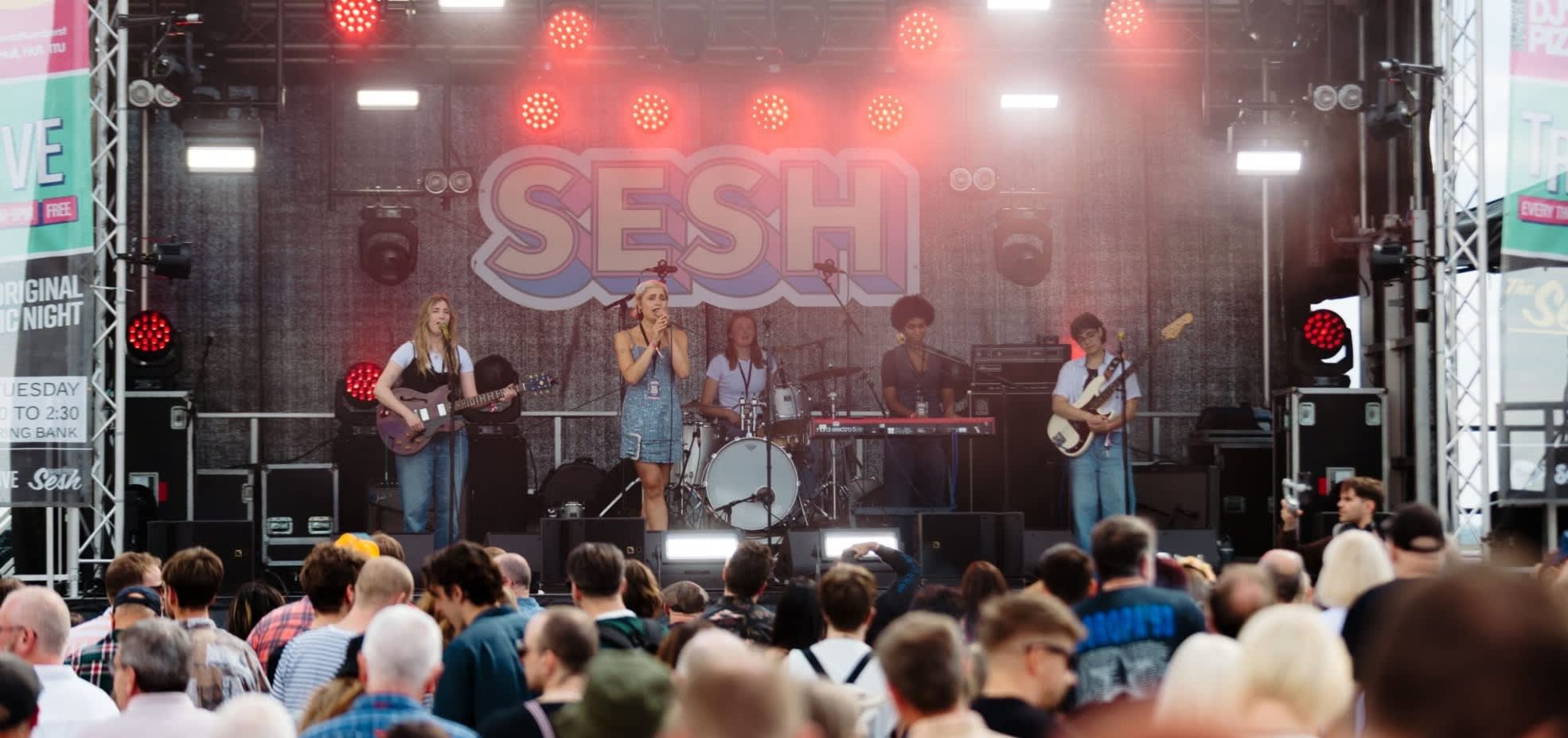 A band sings and plays instruments on stage at Humber Street Sesh to a crowd of people.