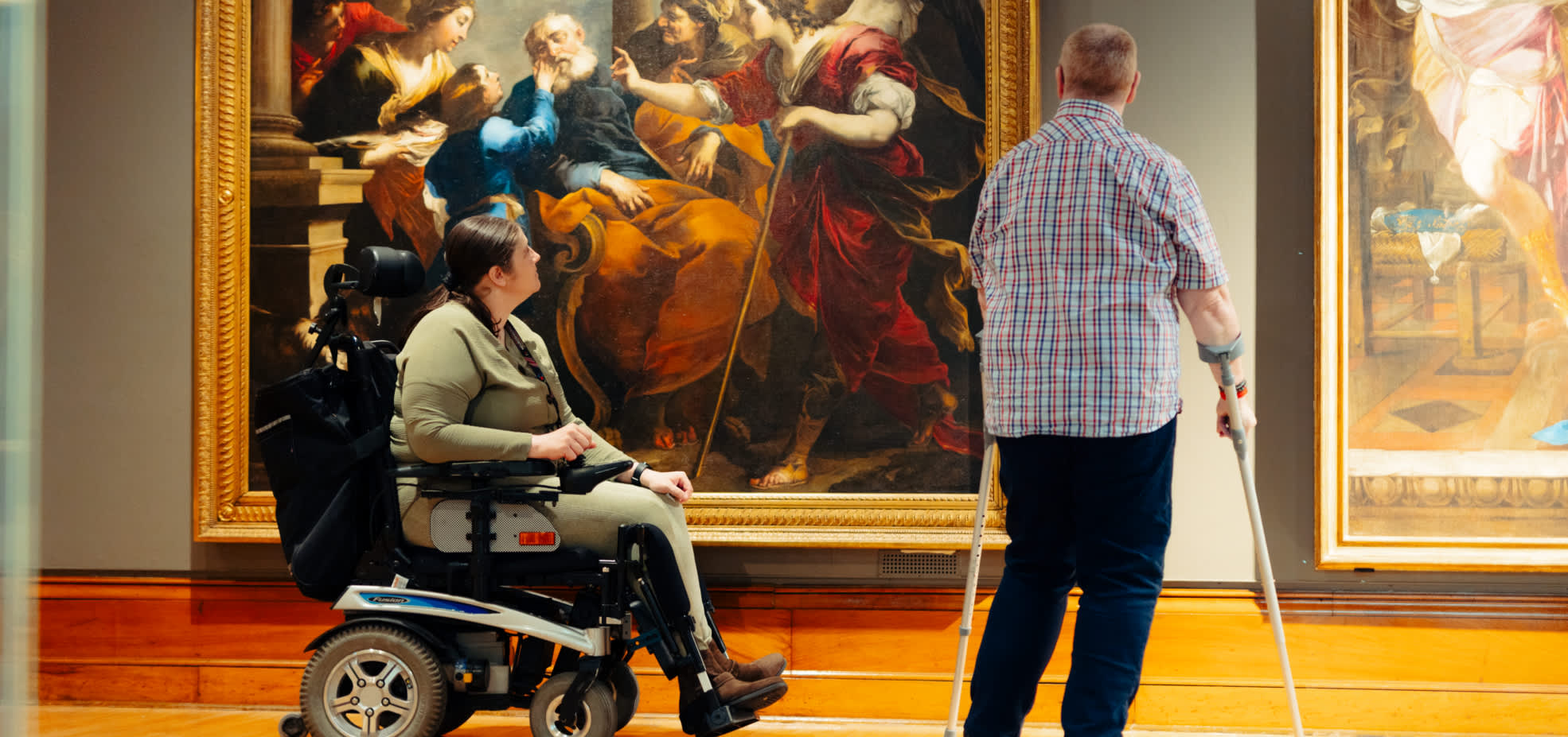 A powered wheelchair user and another visitor with a mobility aid admire the  large classical painting 'Tobias Healing the Blind Tobit' (c.1650) by Valerio Castello in the spacious, accessible Ferens art gallery Hull .