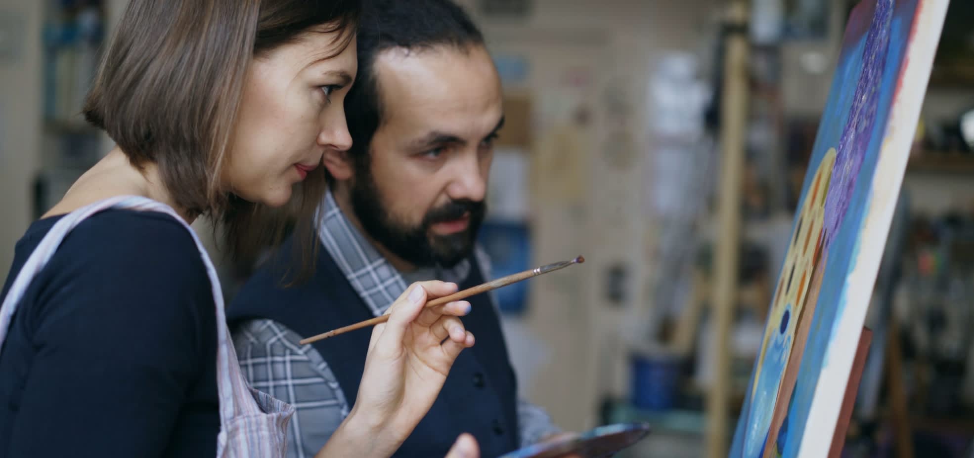 Two artists stand in front of an easel gazing at the painting, mid discussion.