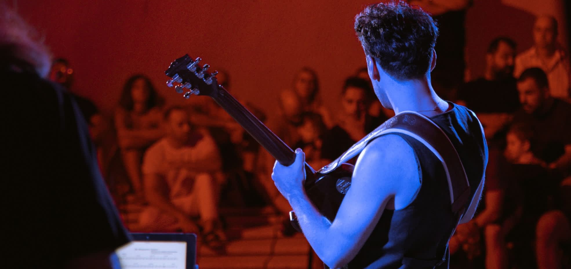 A man plays an electric guitar to an audience. He faces away from the camera and is illuminated by blue stagelights.