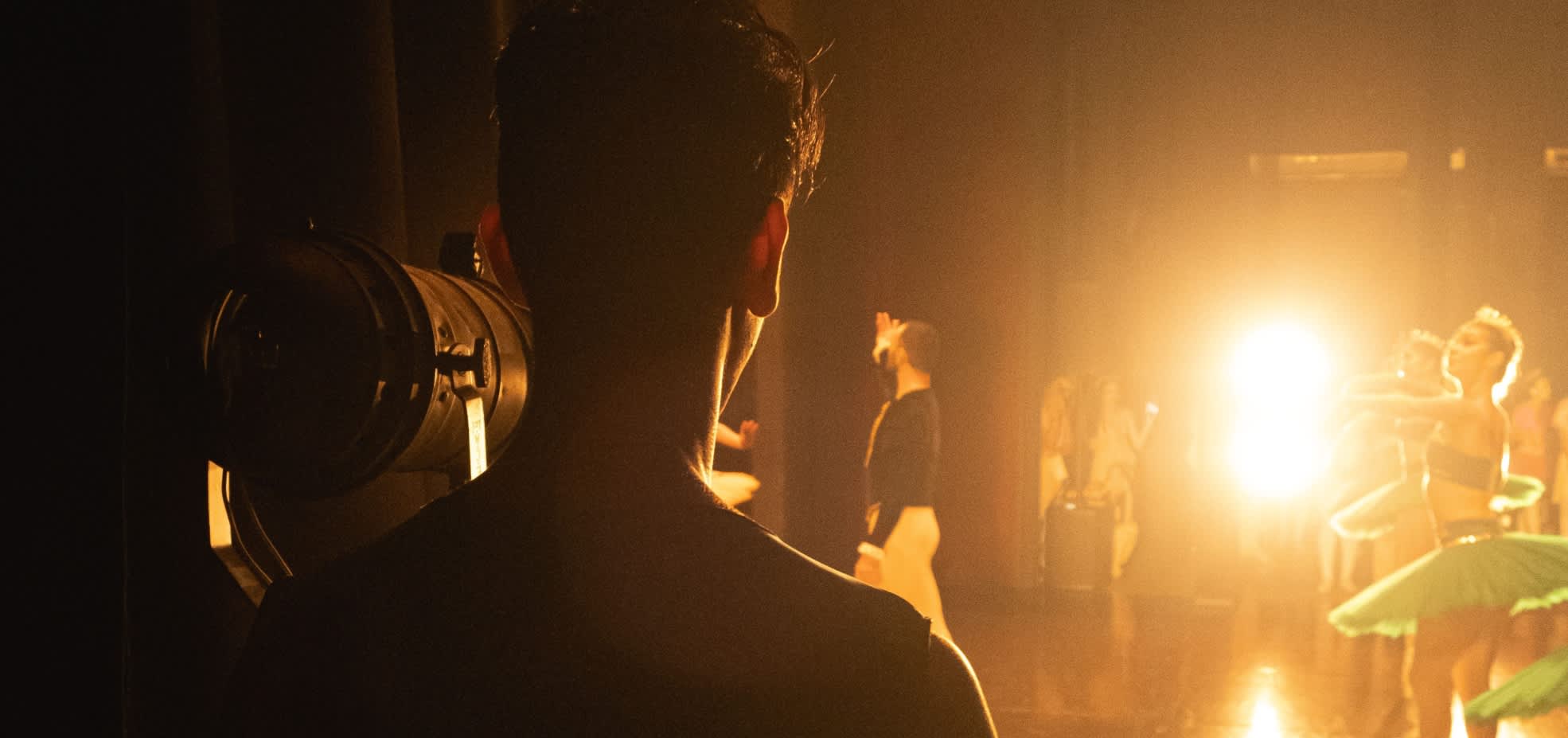 A man stands behind the curtains of a stage waiting to go on whilst ballet dancers are on stage. The spotlight shines towards the man as he faces with his back to the viewer.