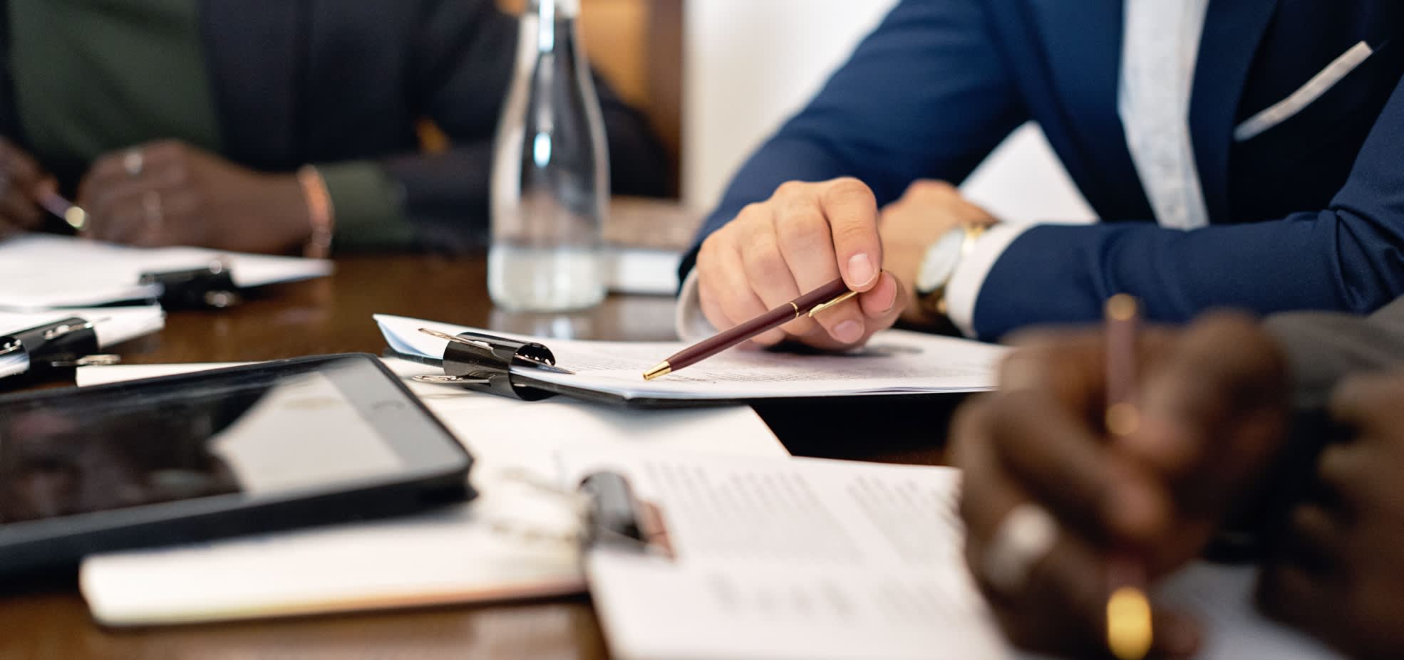 A close up of hands on a table during a meeting. There's paper, a water bottle, calculator and pens on the table.