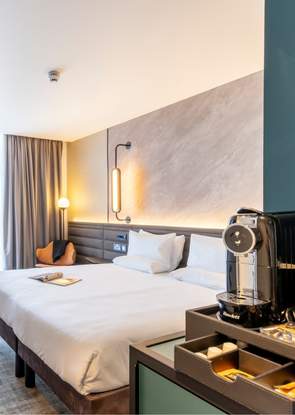 An extra large white bed in a spacious, airy room. The walls are a grey marble on one side and a deep green on the other. To the right is a special coffee machine and nearby a selection of coffee pods.
