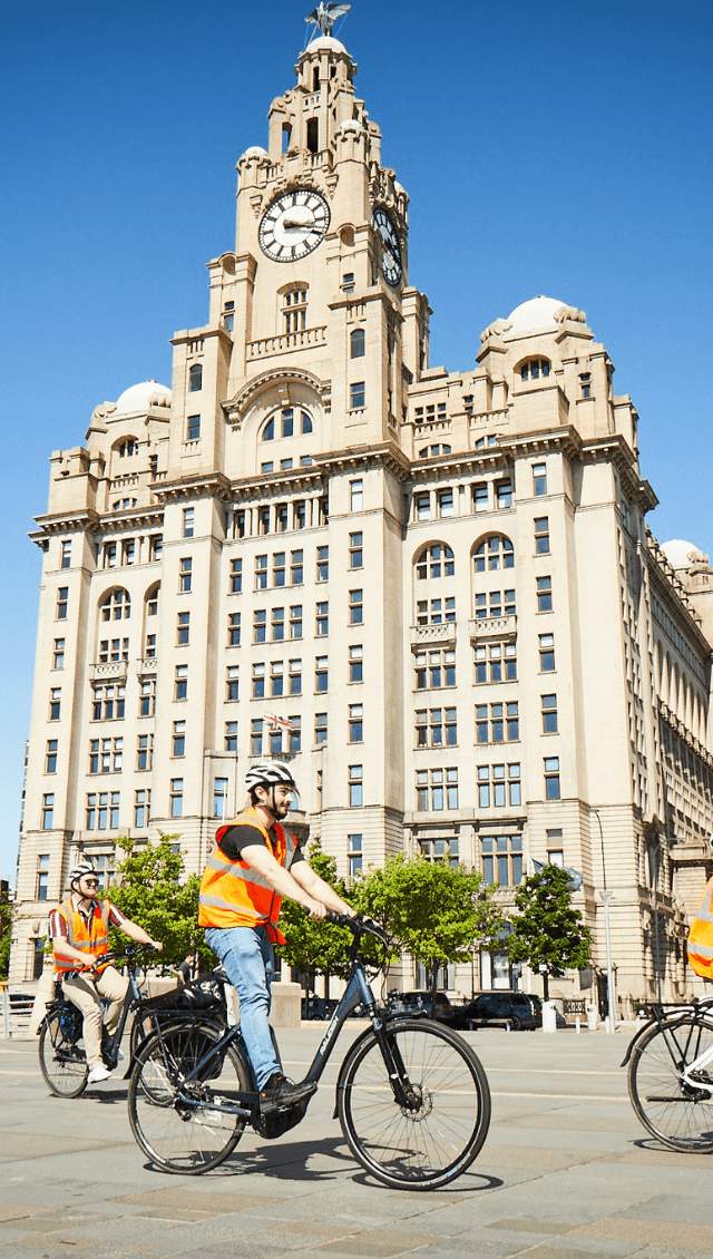 People wearing orange high vis jackets and helmets ride electric bikes in front of the Liver Building.