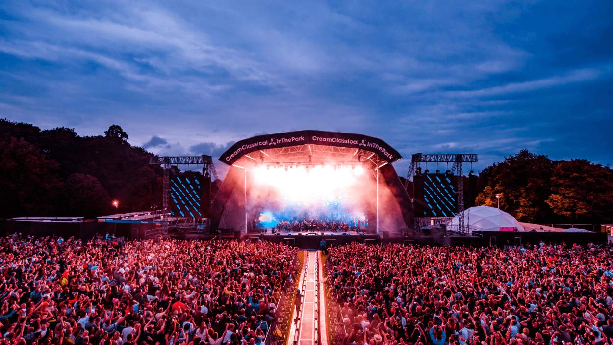 A huge stage and crowd at dusk in Sefton Park. There are lots of lights and pyrotechnics on stage.