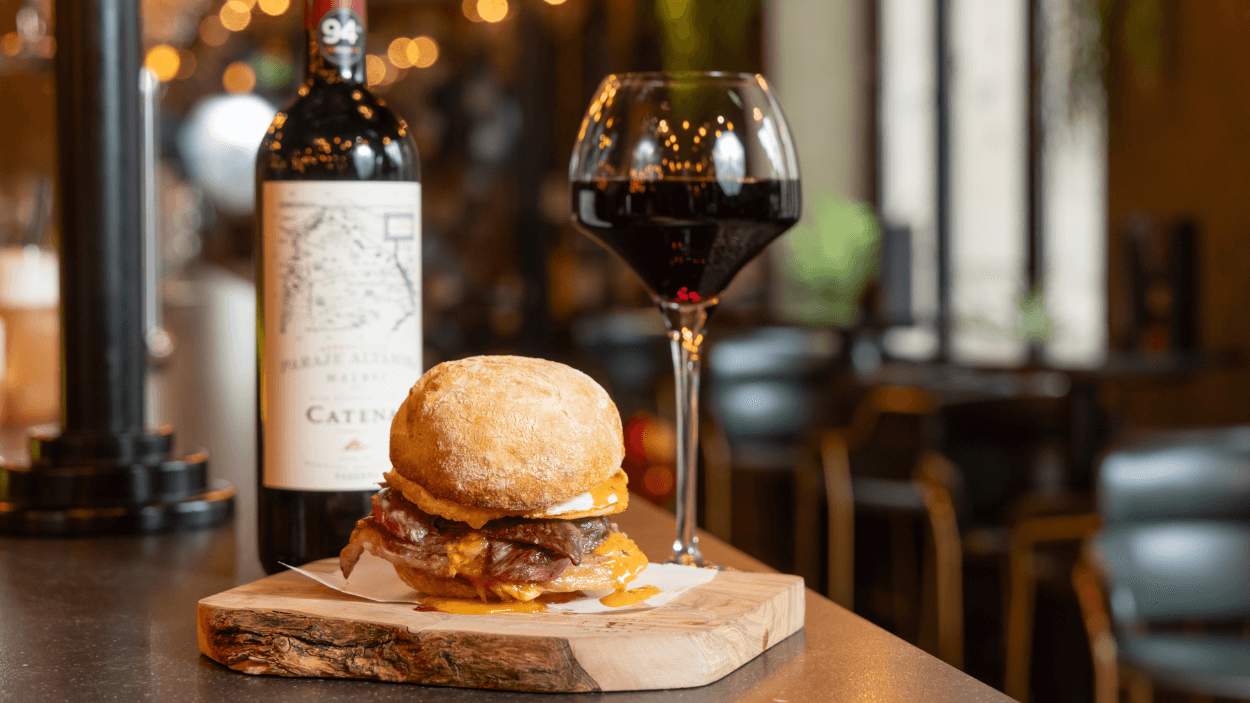 A table with a plate on with a burger on it. There is a bottle of wine in the background and a glass of red wine next to it.