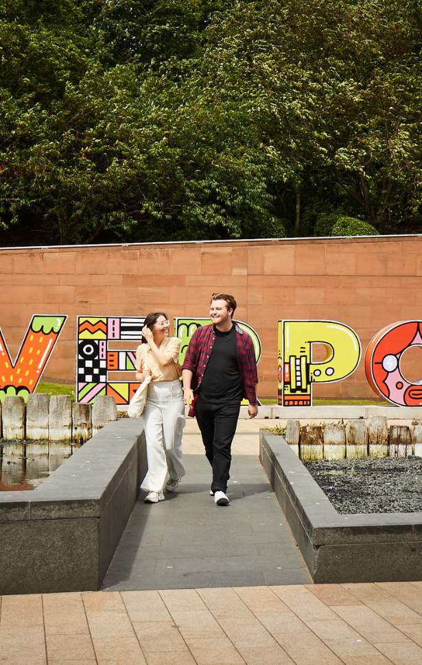 A young female and male walk happily. In the background there is a large 3D letter sign that reads 'Liverpool' and has doodle style colourful drawings decorated on each letter.